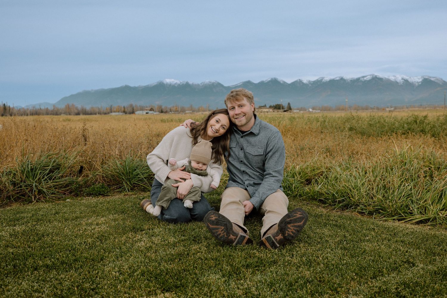 A couple relaxes together in a grassy meadow with mountains in the background on a sunny day.