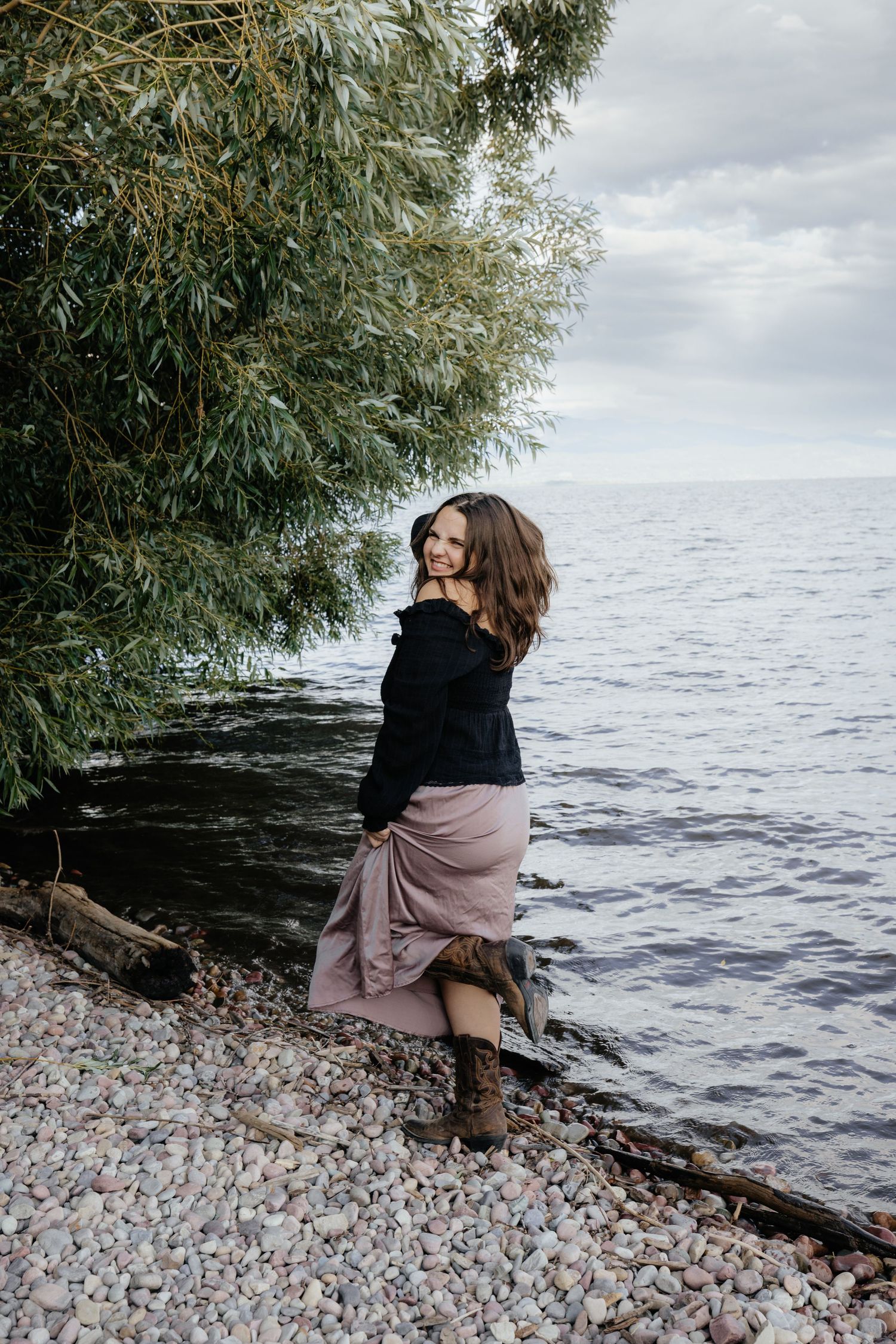 Person in cozy sweater sitting by rocky lakeshore under willow branches.