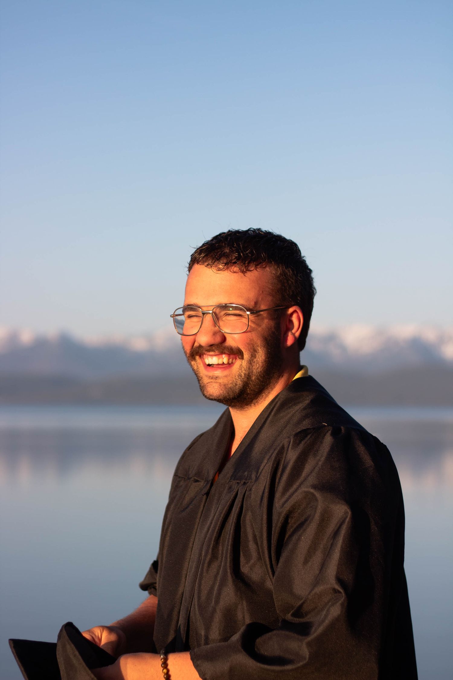 A person in a black jacket smiles warmly against a serene waterfront backdrop during golden hour.