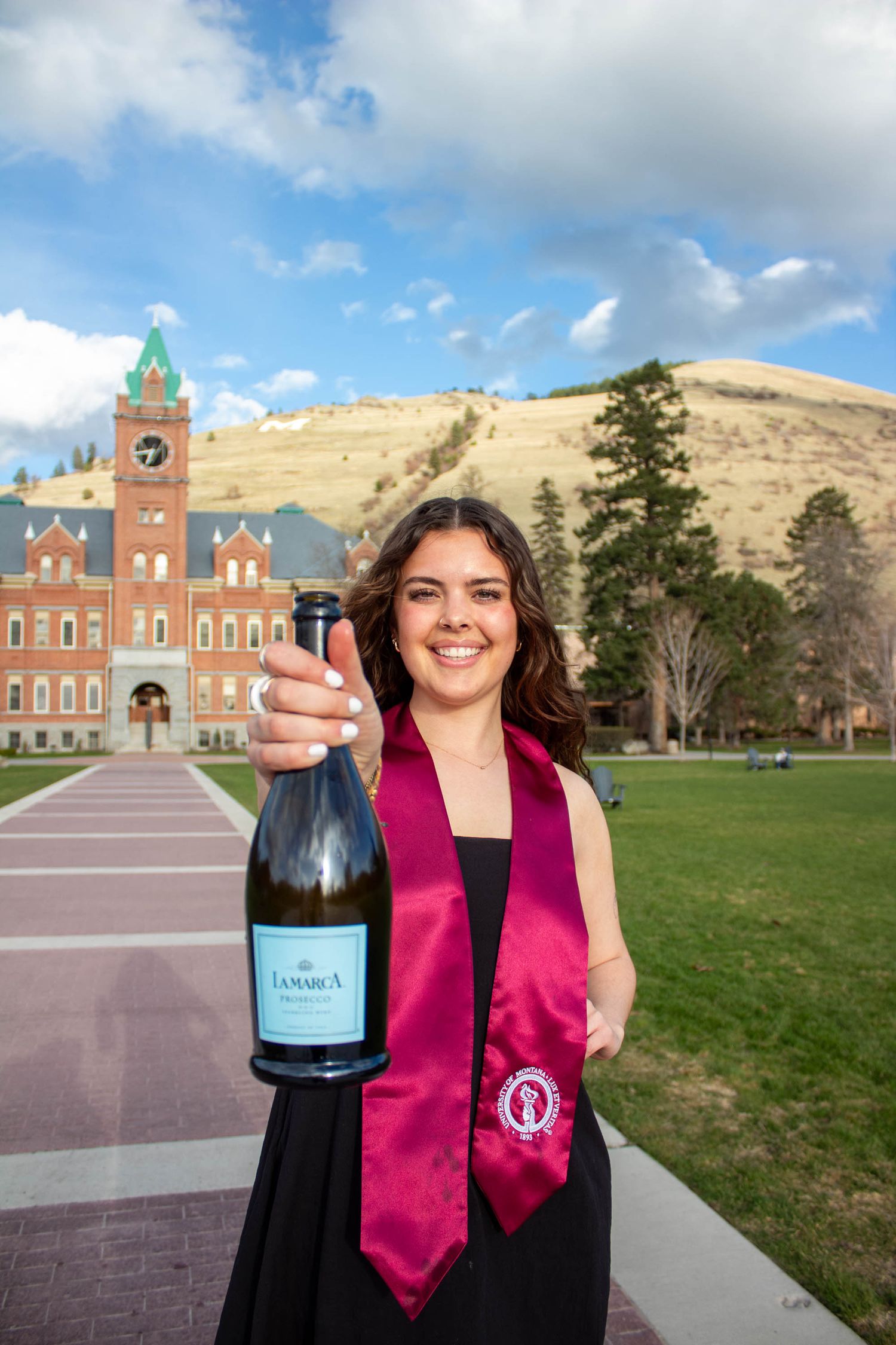 Graduate in academic regalia holding champagne bottle outside historic brick university building.