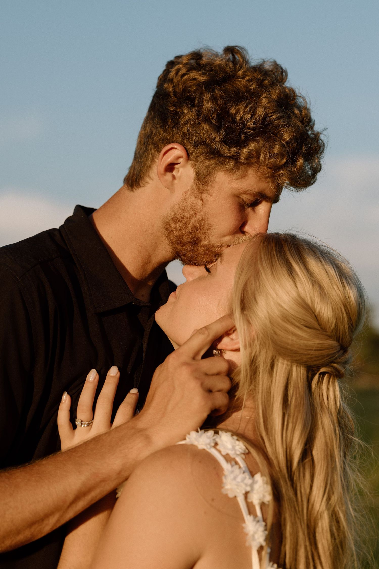 A romantic couple shares an intimate kiss outdoors during what appears to be a wedding photoshoot at sunset.