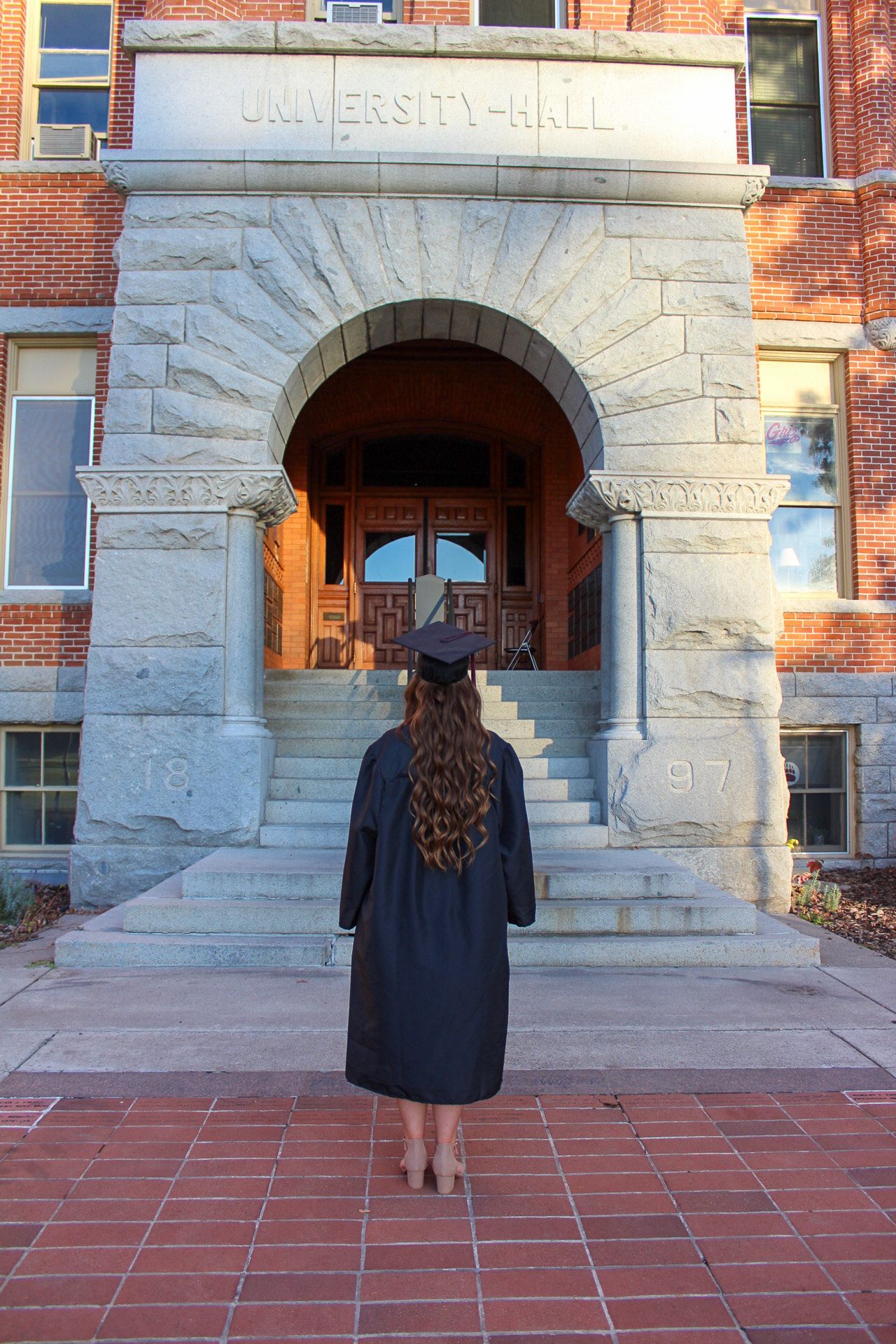 Graduate stands in academic regalia in front of ornate stone university building entrance with brick walls.