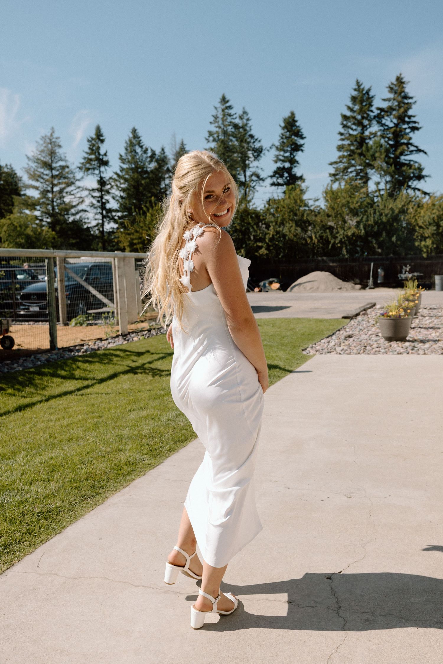 Woman in elegant white dress poses on sunny patio with evergreen trees in background.