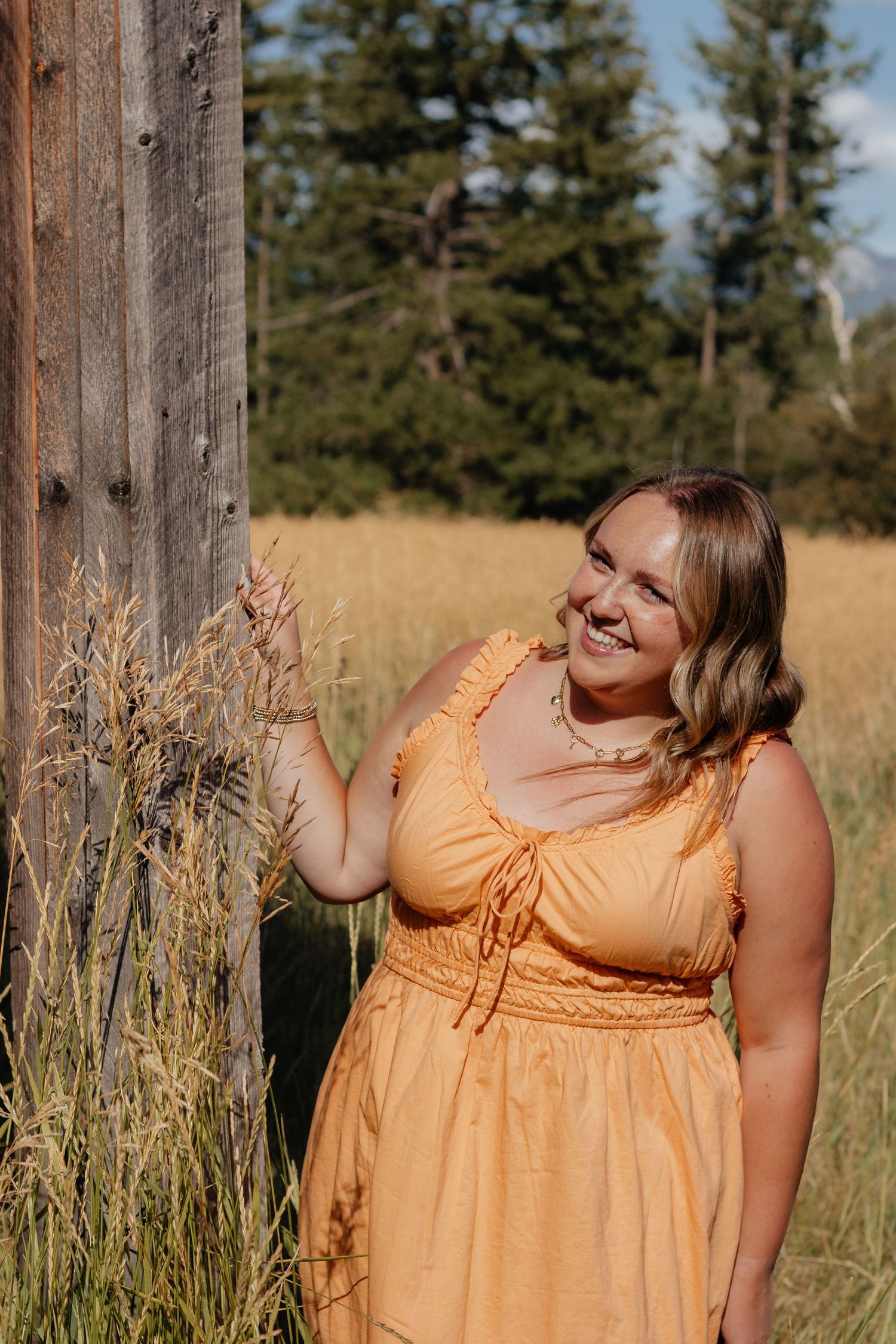 A person in a peach sundress poses in a field with evergreen trees and mountains in the background.