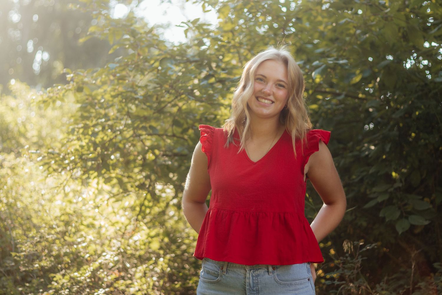 Woman in red top and jeans poses outdoors in natural sunlit setting with trees and foliage in background.