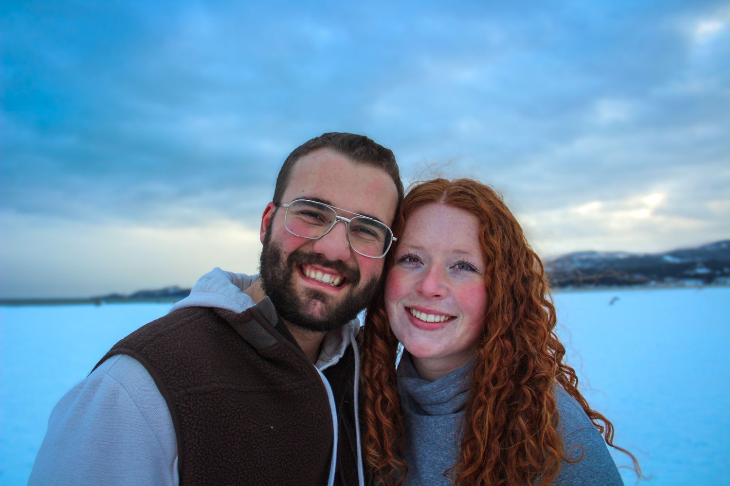 A happy couple poses for a selfie against a snowy winter landscape with blue sky at dusk.