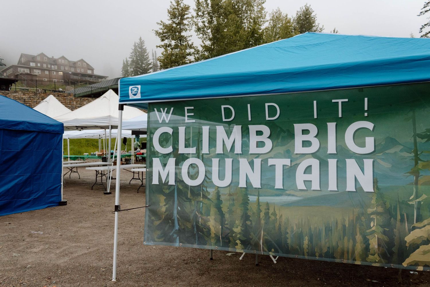 Banner at outdoor event reads 'WE DID IT! CLIMB BIG MOUNTAIN' against scenic mountain backdrop.