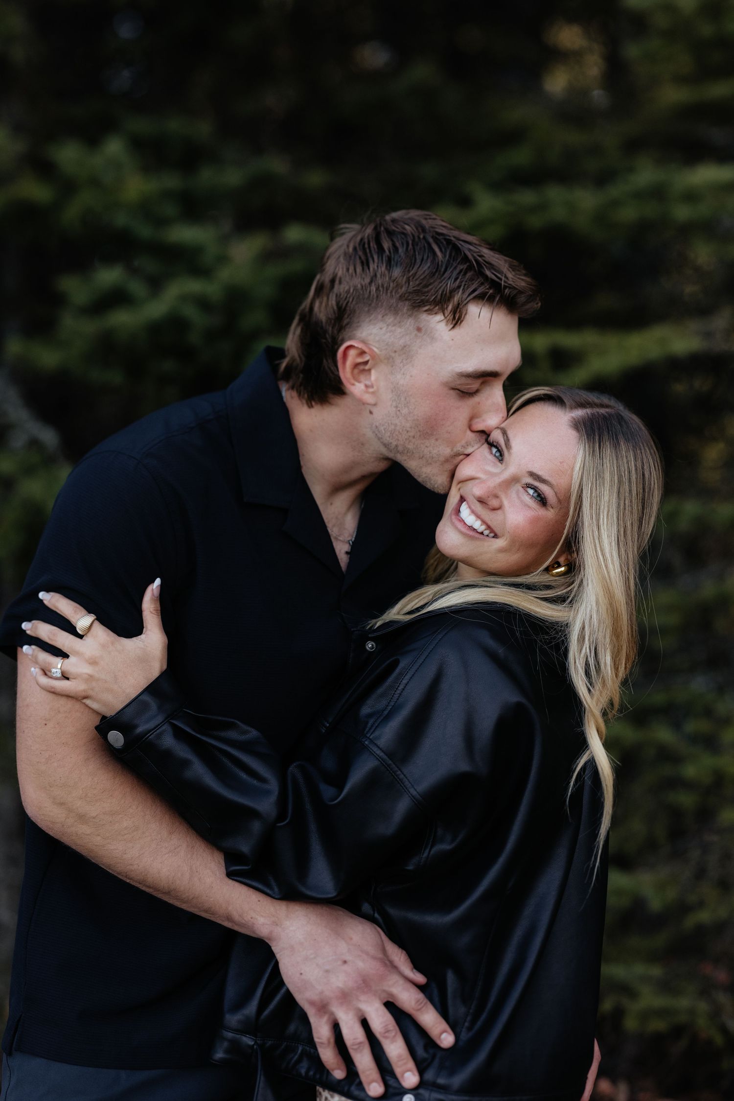 A couple in black outfits embracing outdoors, man kissing woman's cheek as she smiles, with evergreen trees behind.
