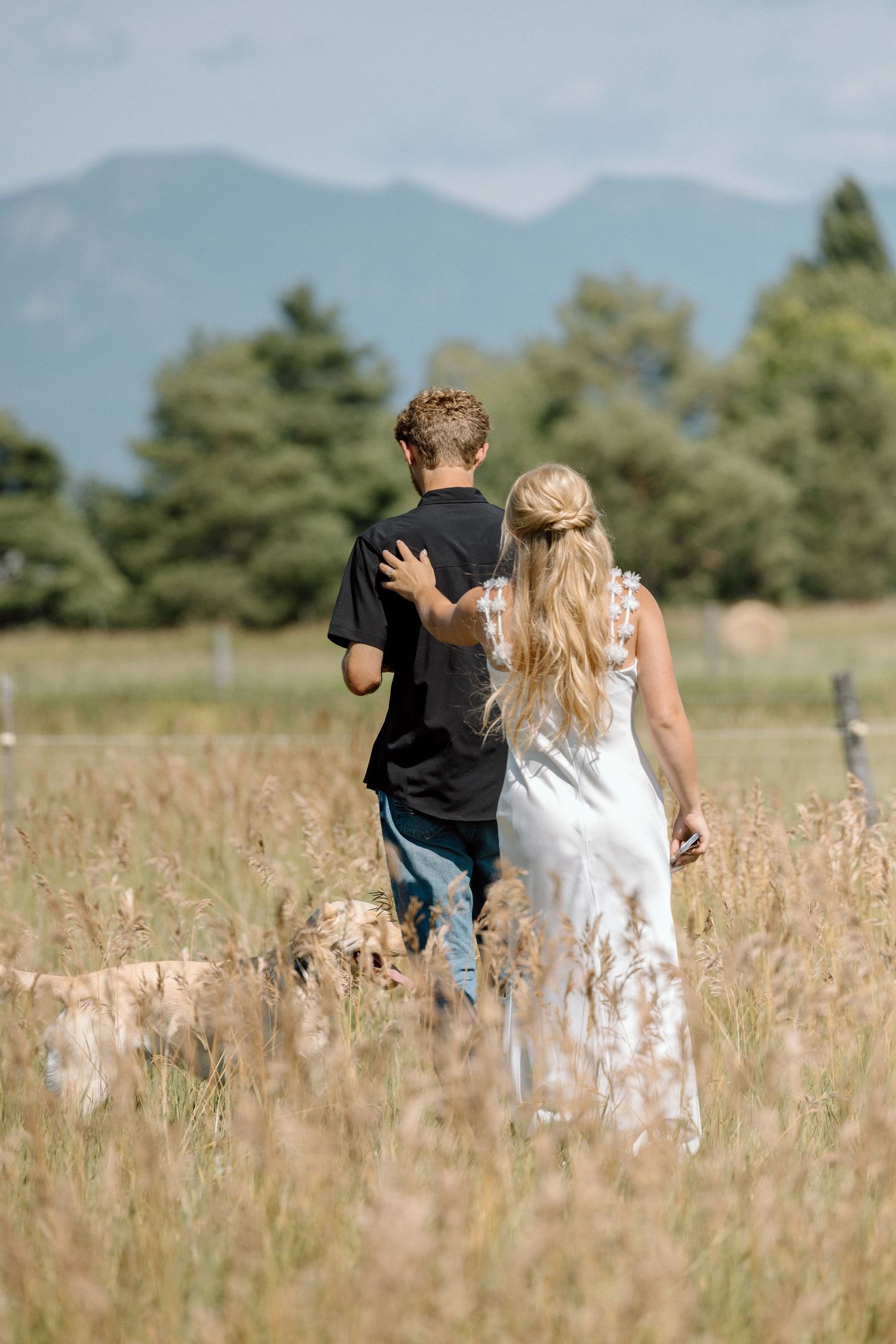 A couple walks through a tall grass meadow with mountains and evergreen trees in the background.
