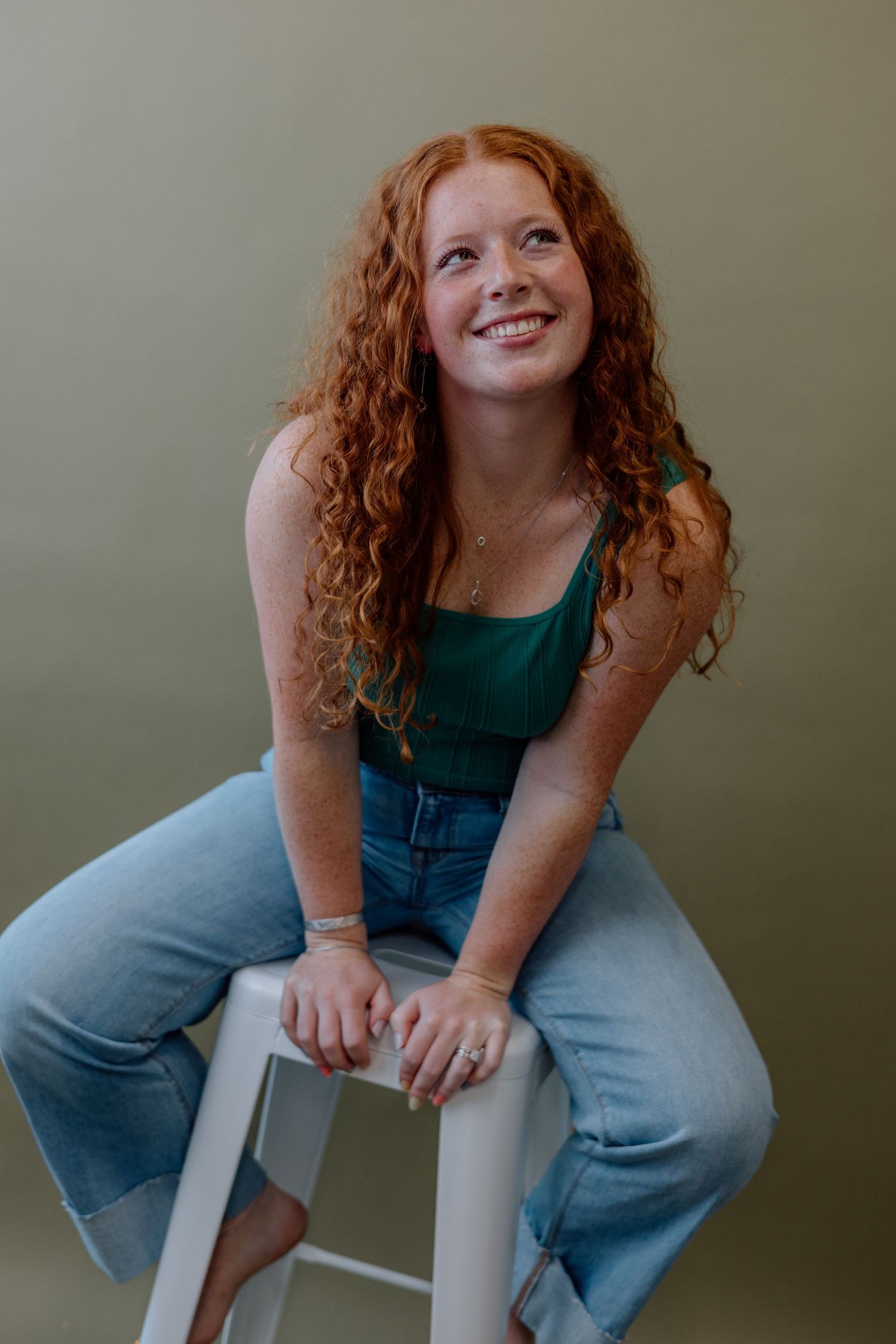 A person with curly red hair wearing denim overalls and a green tank top sits casually on a white chair.