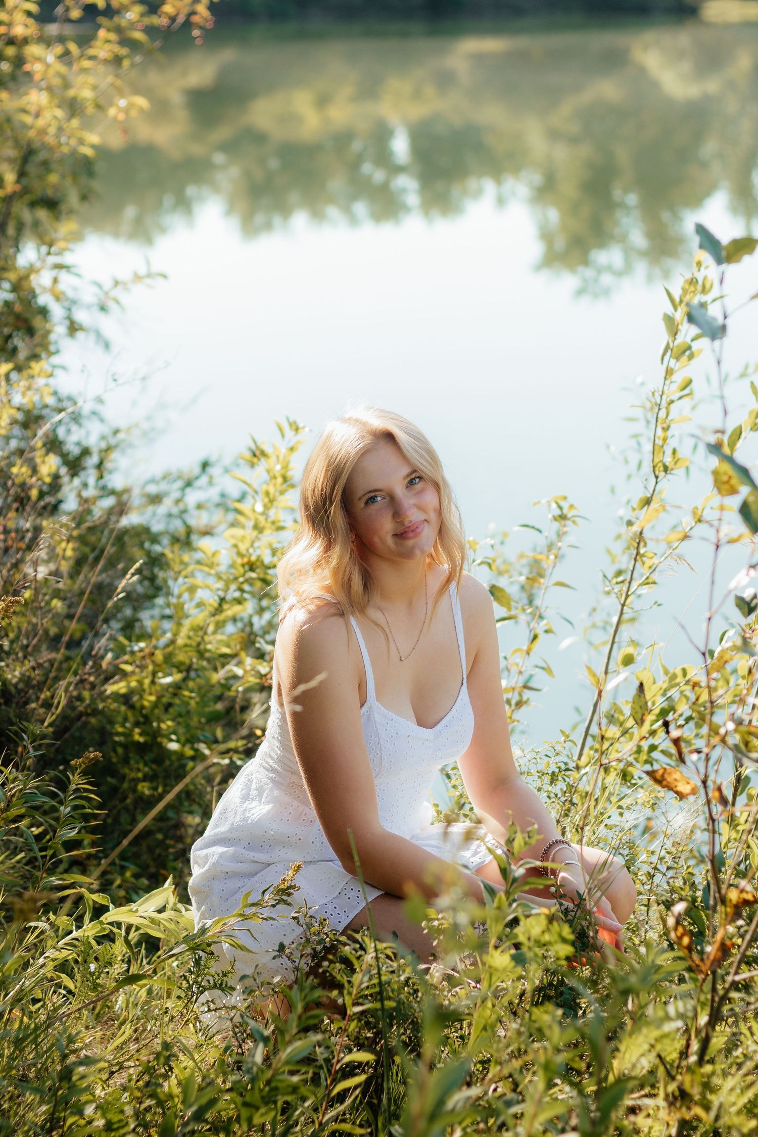 A person in a white dress sits among wildflowers by a tranquil lake on a sunny summer afternoon.