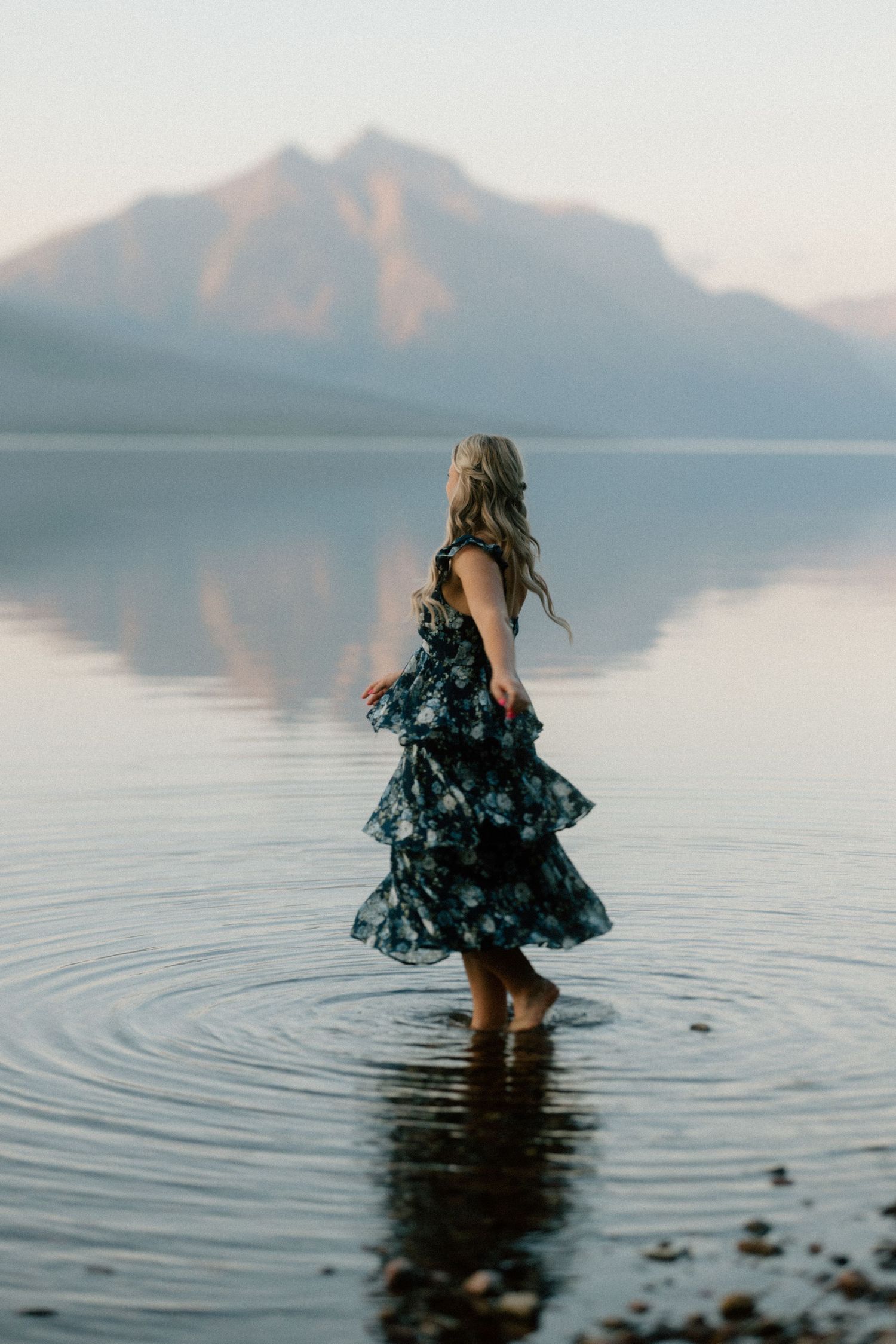 A figure in a floral dress wades deeper into a serene mountain lake at sunset creating rippling reflections in the water.