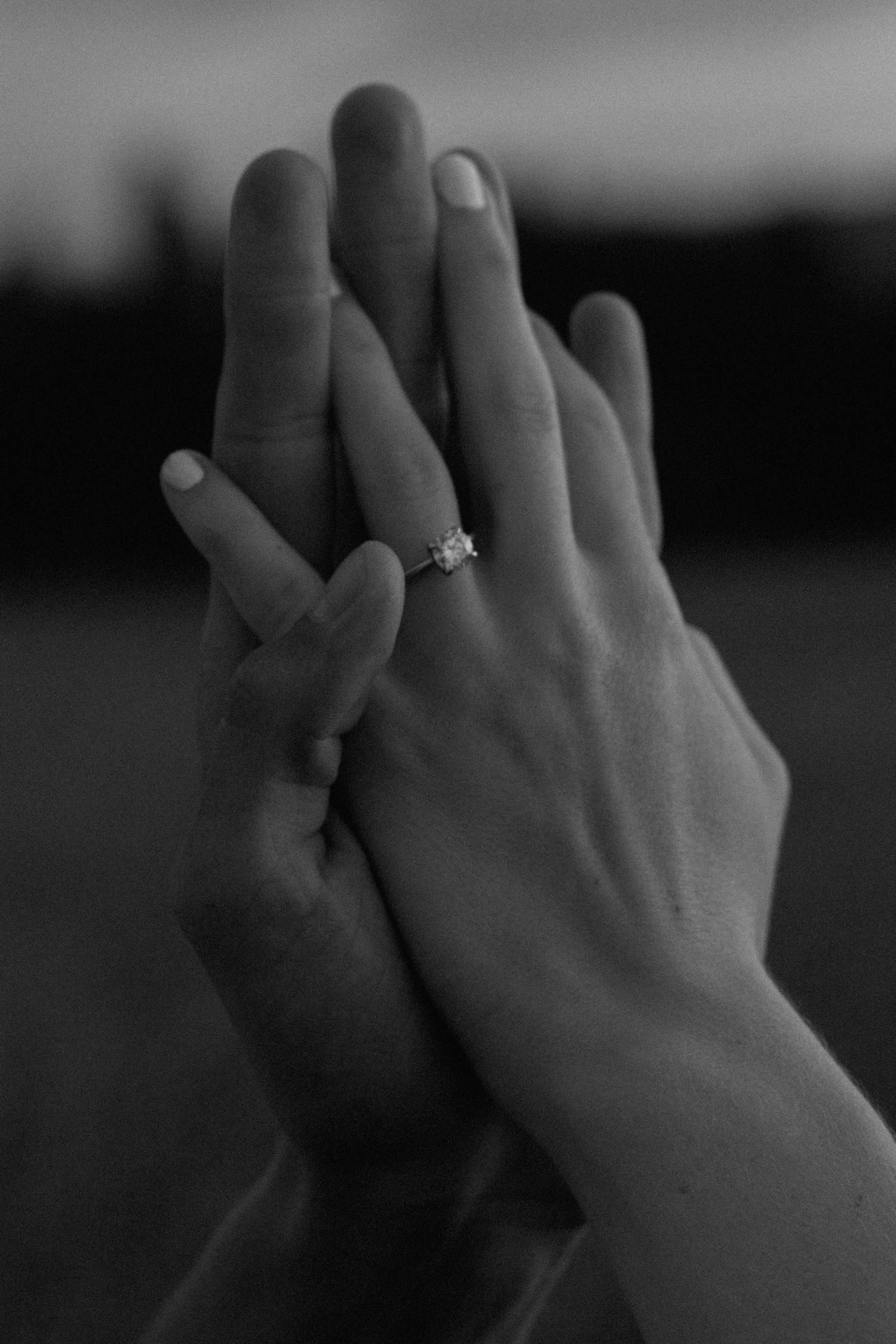 Two hands intertwined in a close-up black and white shot showing an engagement ring.
