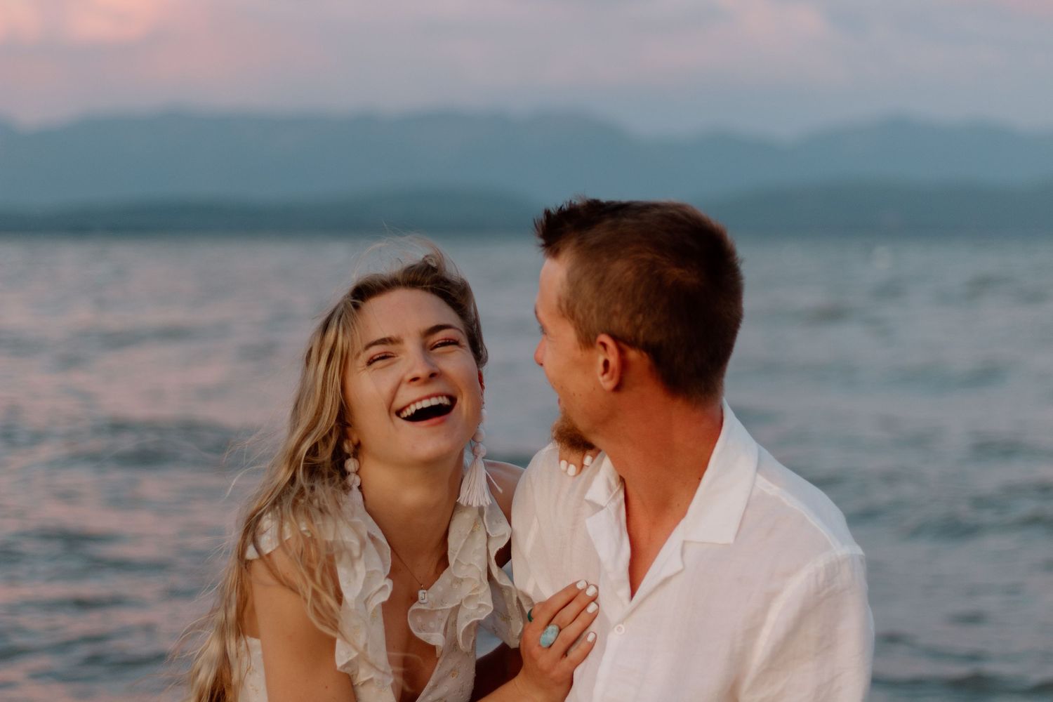 Romantic couple embraces and laughs together during sunset photoshoot by the ocean.