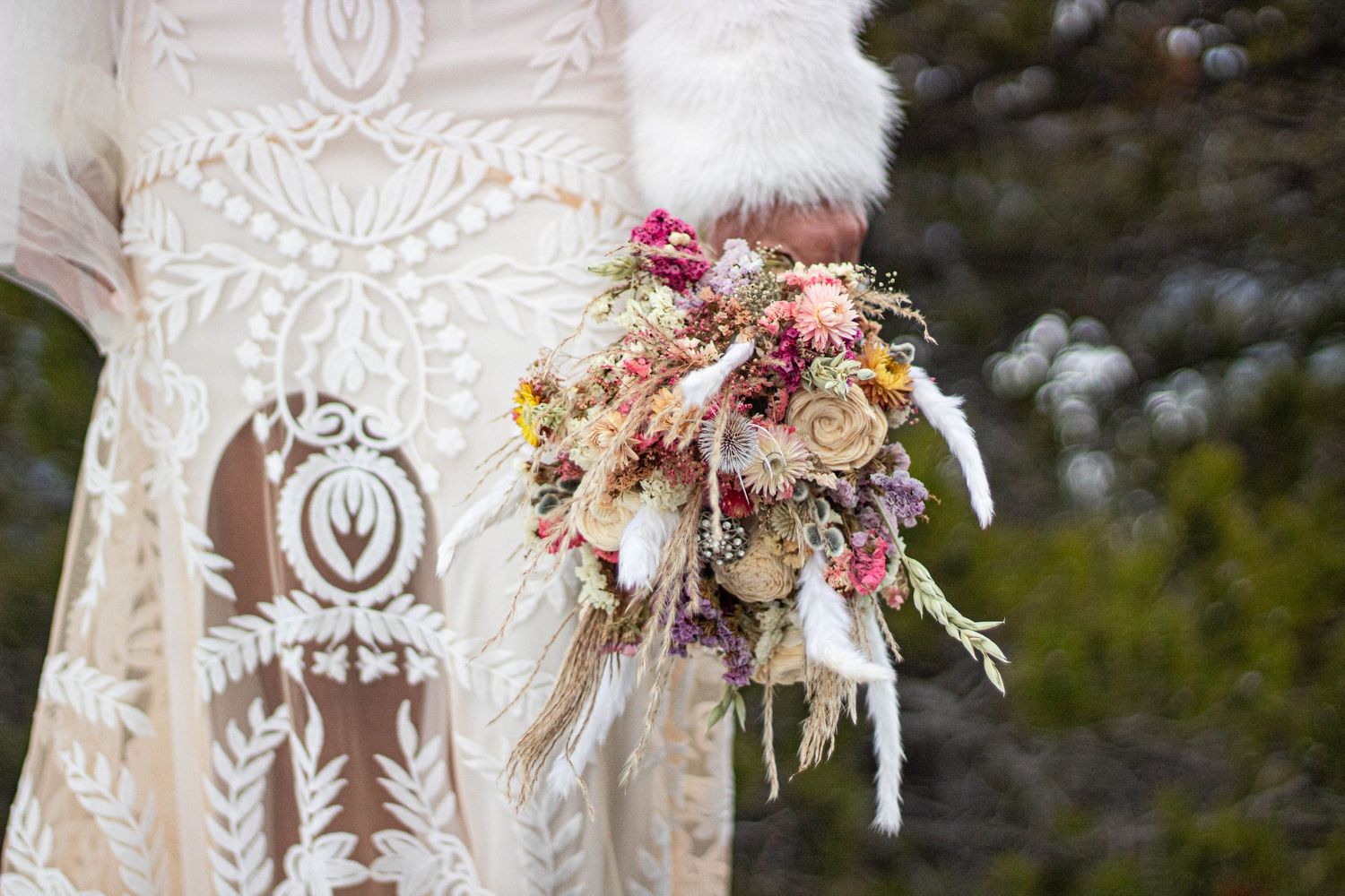 A delicate dried flower bouquet with pink and cream blooms held against a white lace wedding dress.