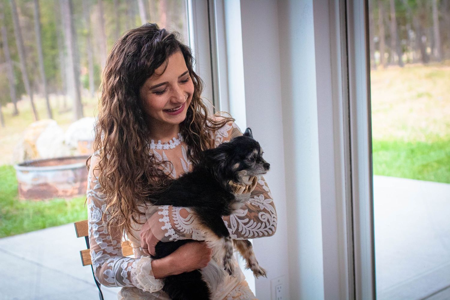 A person in a white lace dress holds a small black and white cat while standing by a window.