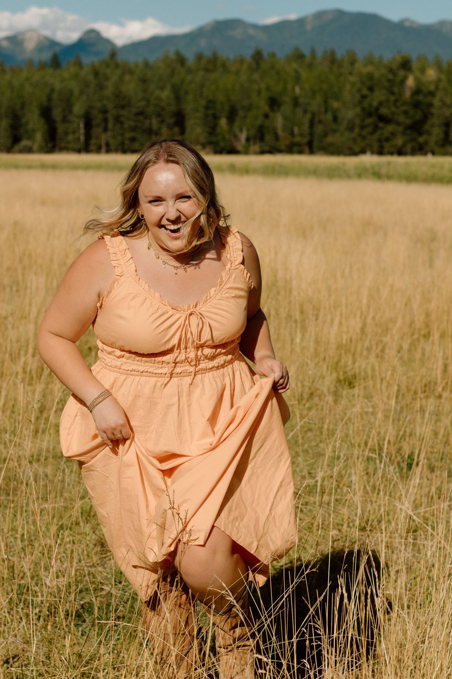 A person in a peach-colored dress poses playfully in a golden field with mountains and evergreen forest in the background.