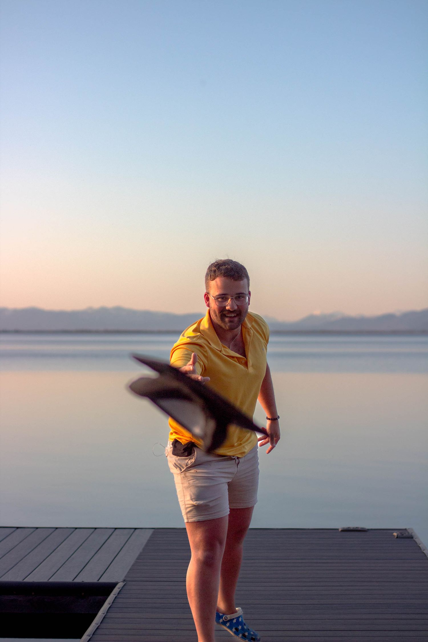 Person in yellow shirt and white shorts makes playful poses on a wooden dock at sunset over calm waters.