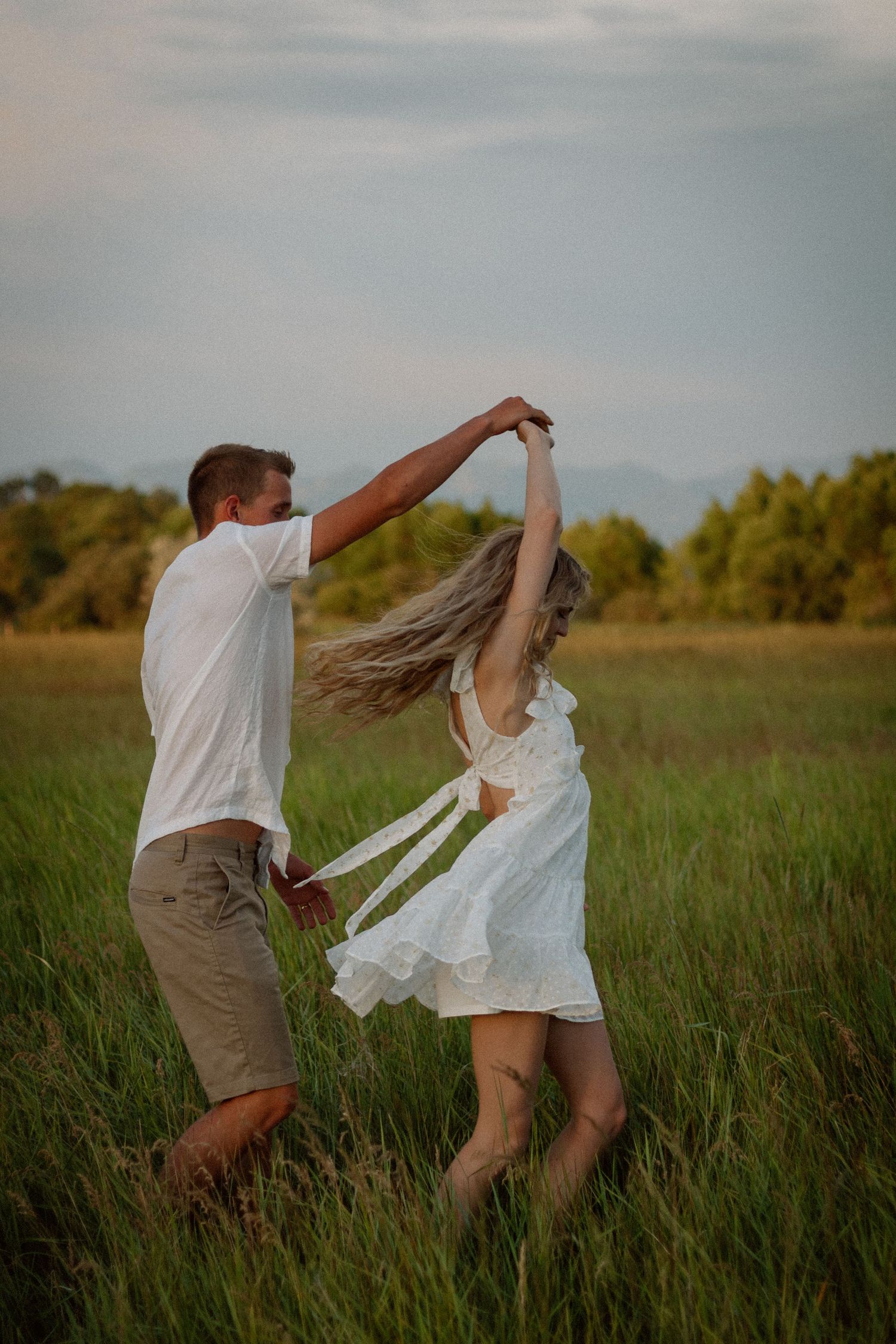 A couple dances together in a grassy field at sunset, with the woman's white dress flowing in the breeze.