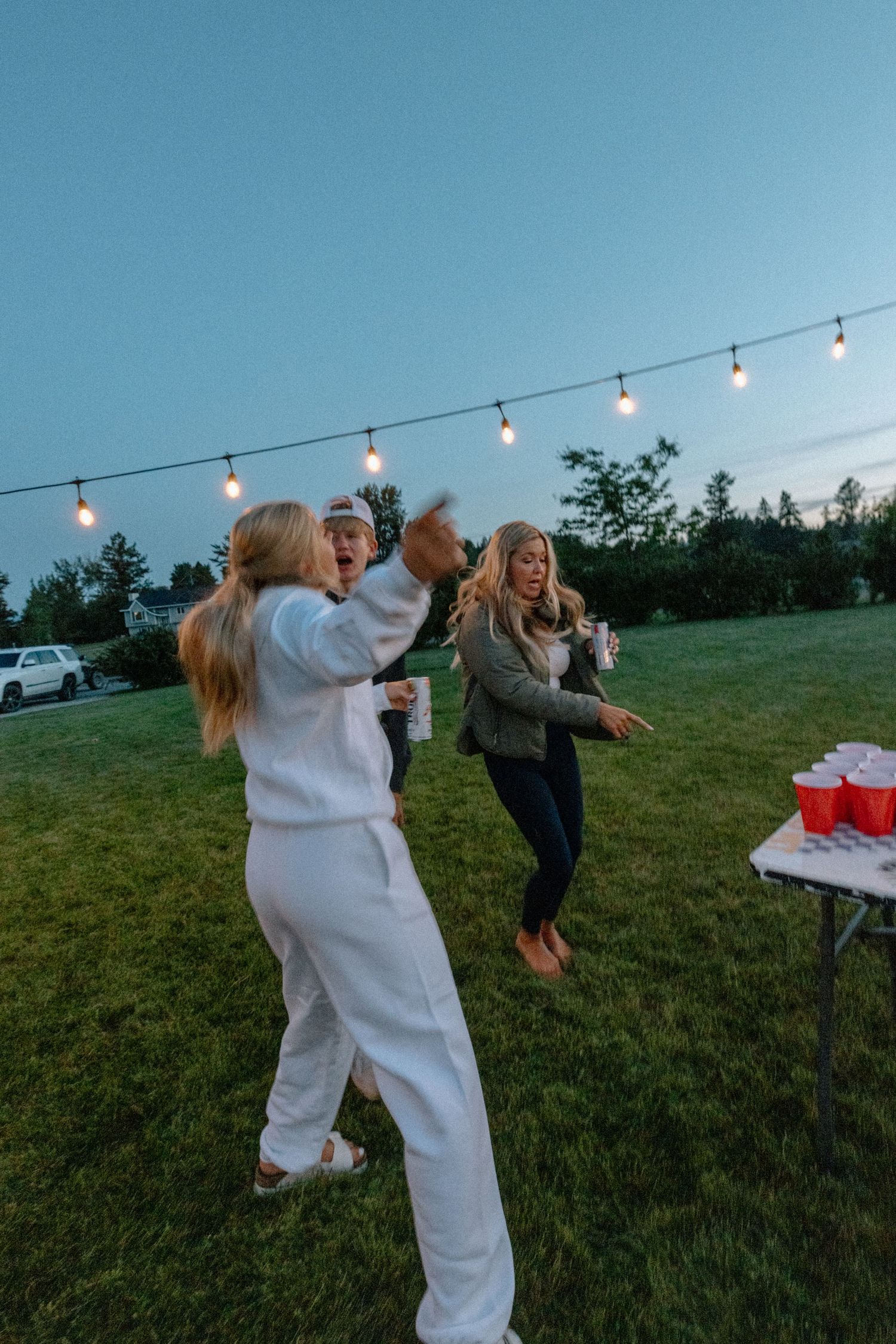 People playing an outdoor party game with string lights overhead in a grassy field at dusk.