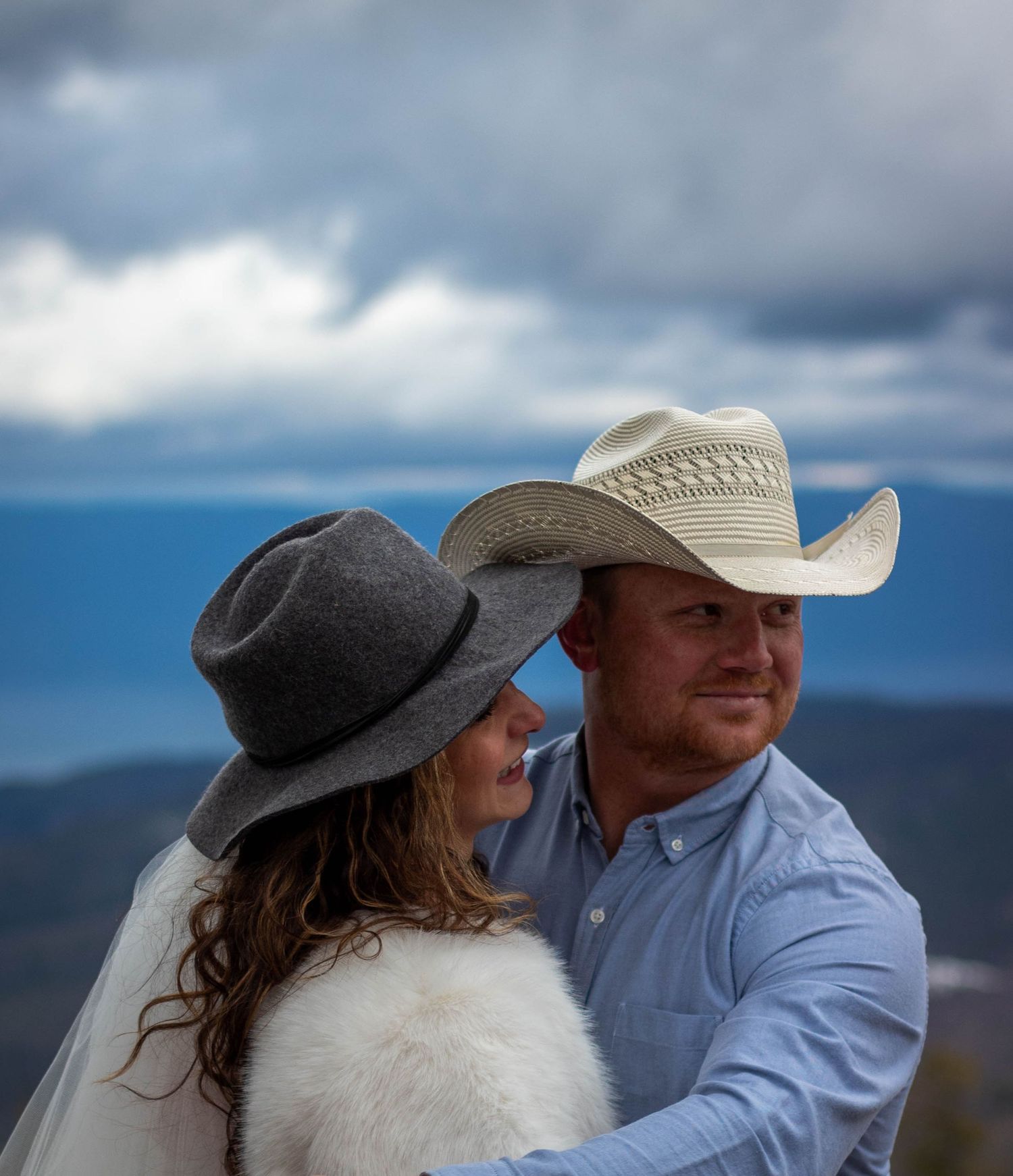 A couple in casual attire share an intimate moment against a dramatic cloudy mountain landscape backdrop.