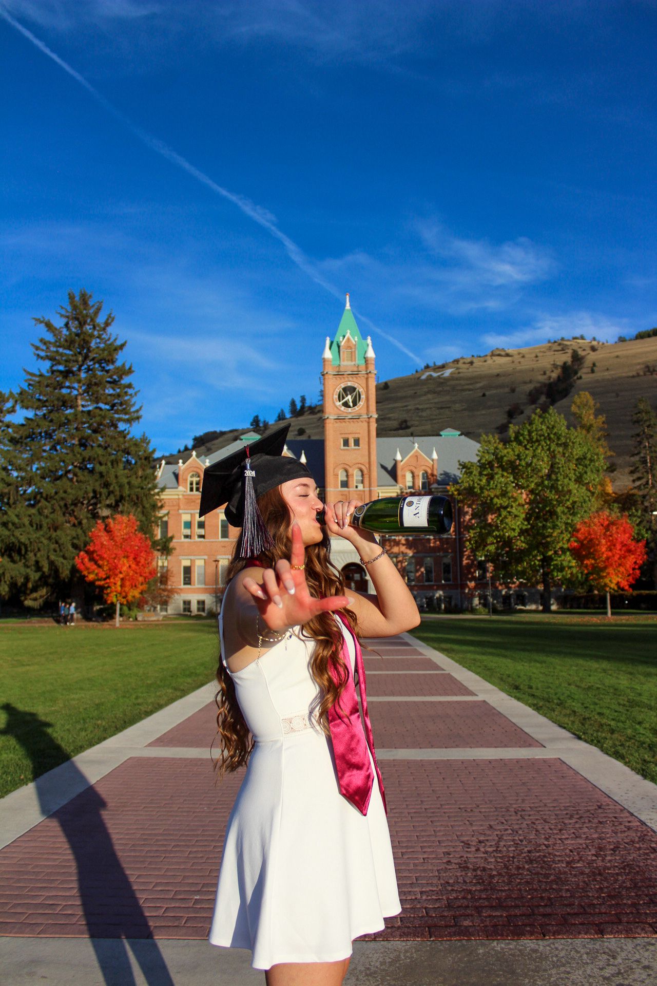 Person in a white dress photographs historic brick building with clock tower against blue autumn sky on campus.