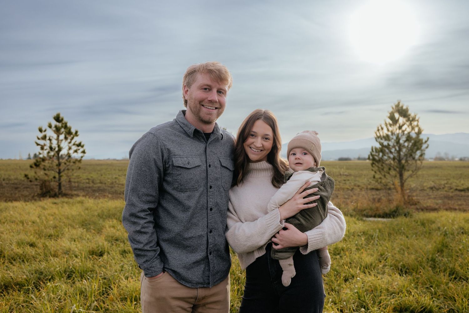 A family poses together on a grassy field with scattered pine trees and cloudy skies in the background.