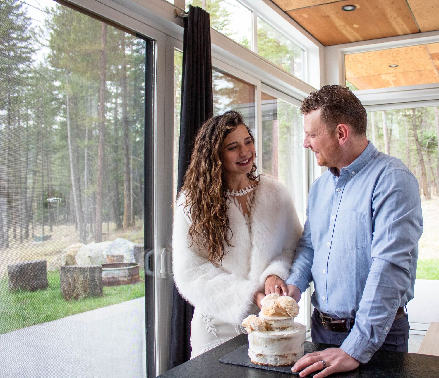 A couple in casual attire cuts their wedding cake together by a large window overlooking a forest landscape.