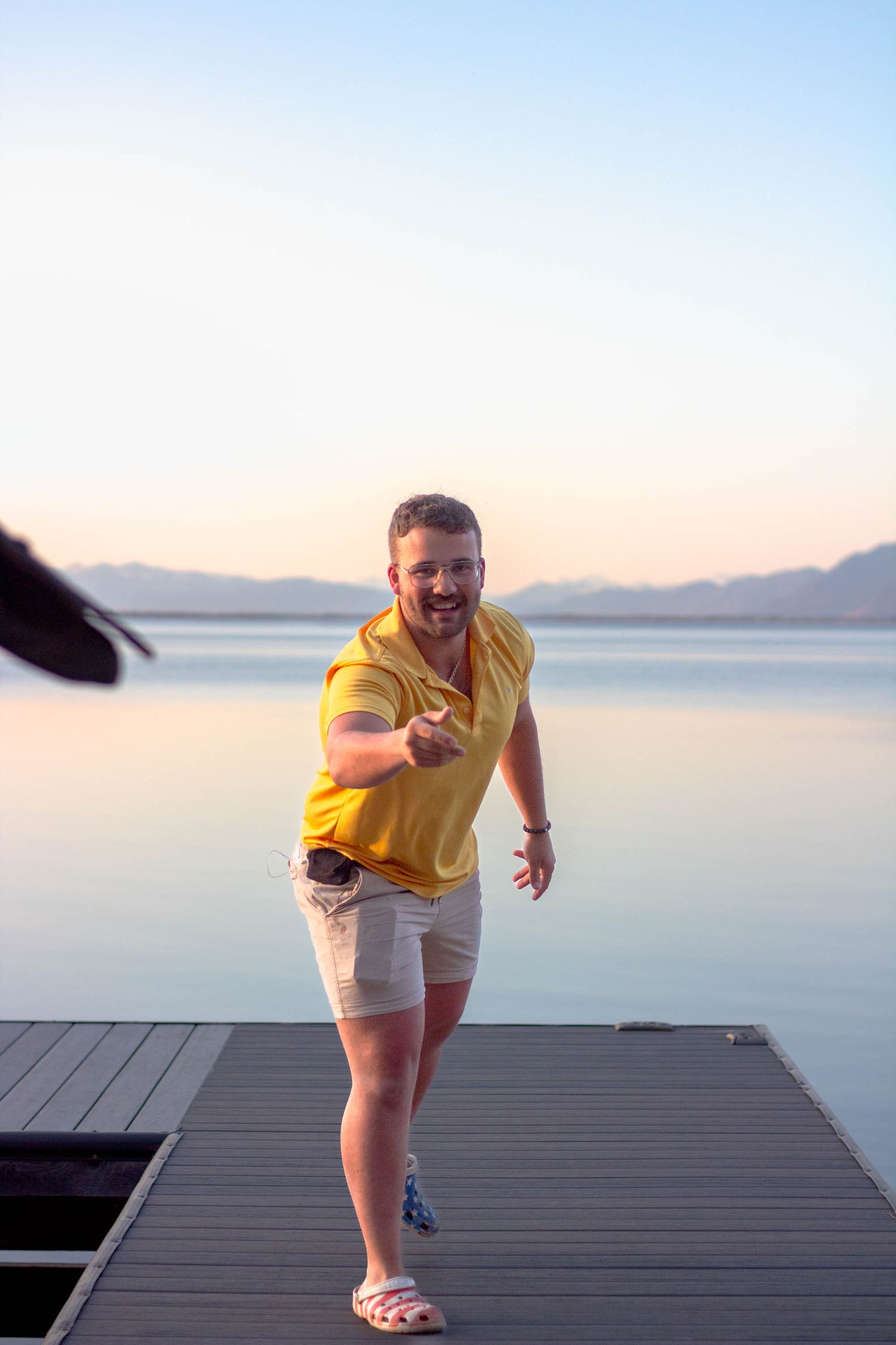 Someone in a yellow shirt and shorts walks playfully across a wooden dock over calm water at sunset.