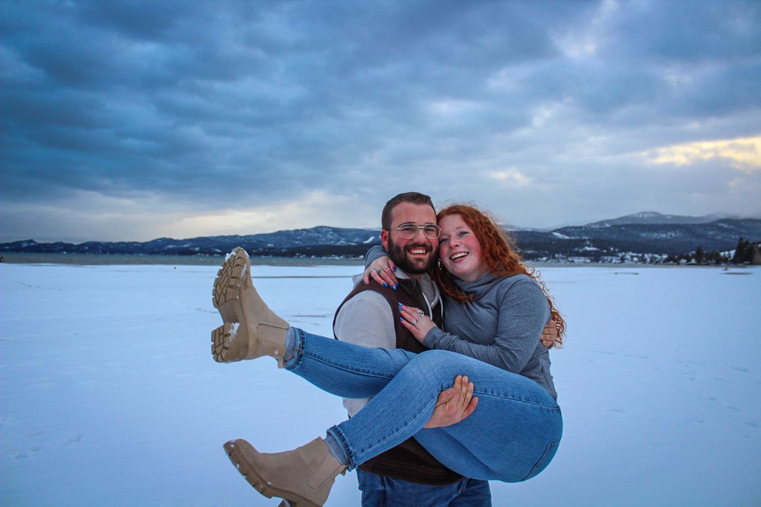 Joyful couple shares intimate moments during winter photoshoot with mountainous background.