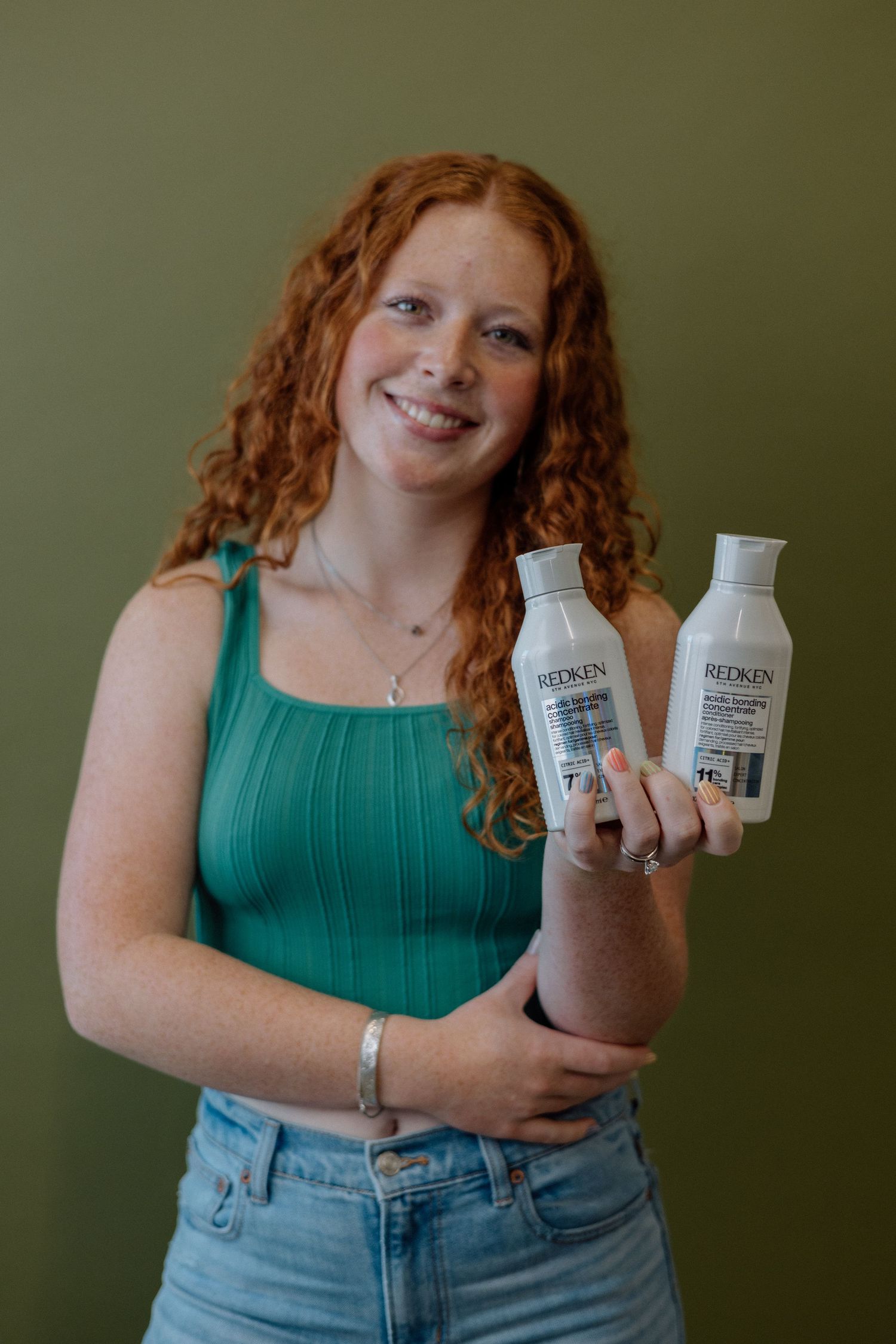 Someone in a teal tank top and jeans poses with milk bottles against a green background.