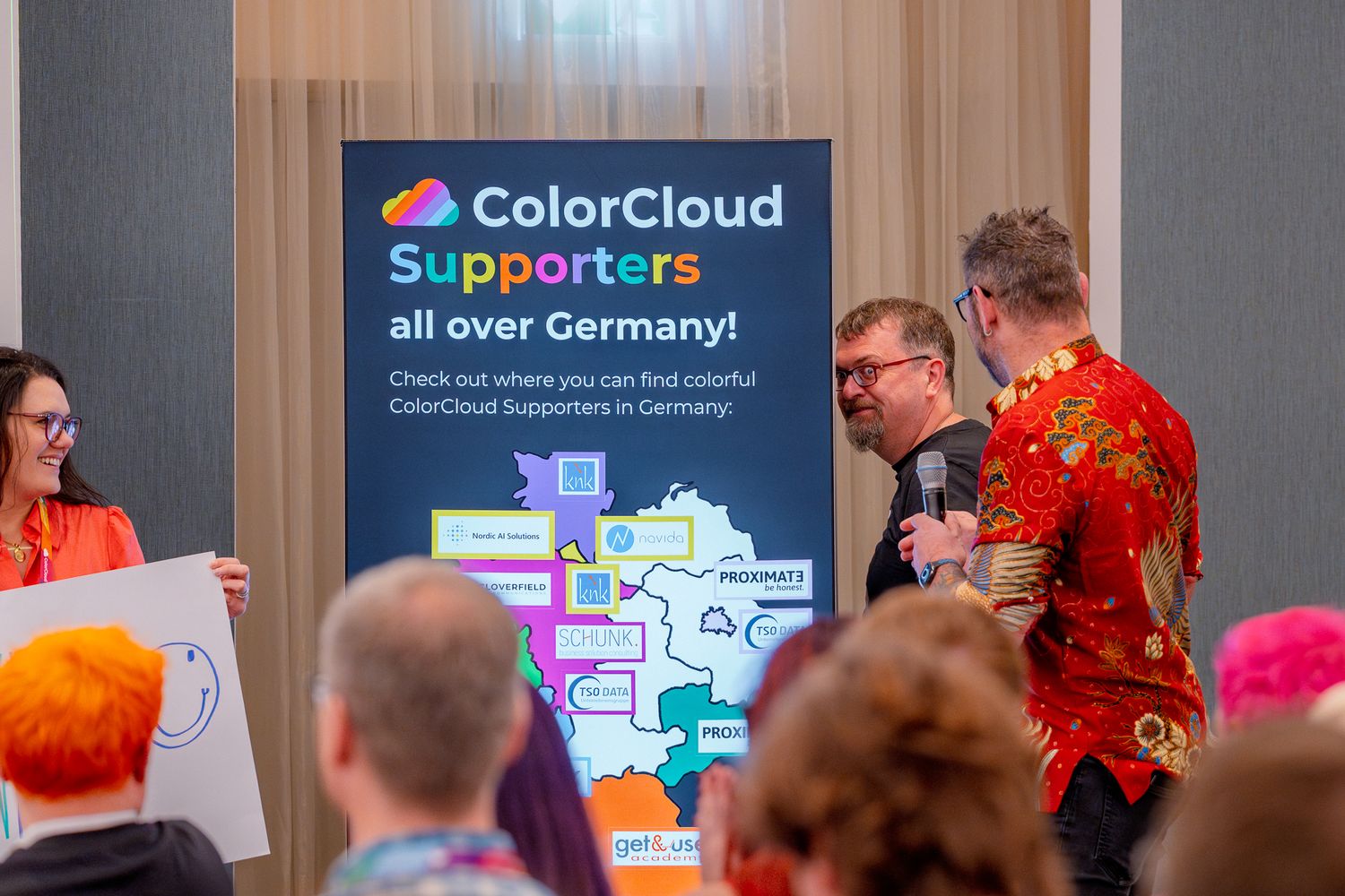 Chris Huntingford führt einen Speaker während der Keynote auf der ColorCloud Konferenz hinter ein großes Branding-Schild