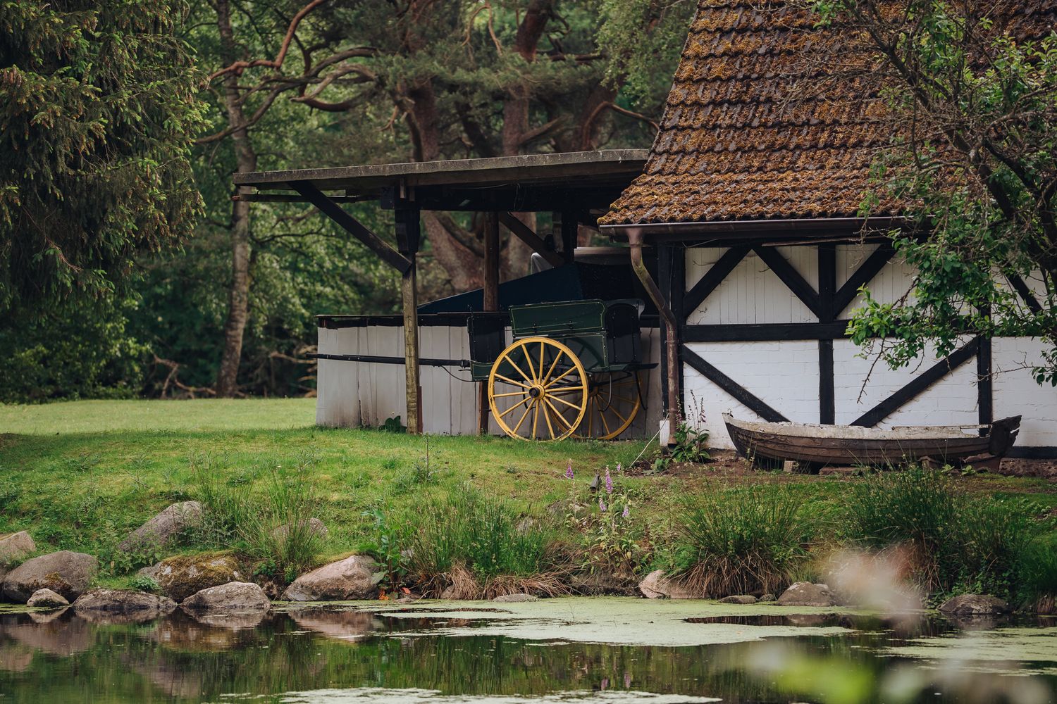 Fachwerk-Nebengebäude mit Kutsche und Holzboot am Ufer des Lütjensees