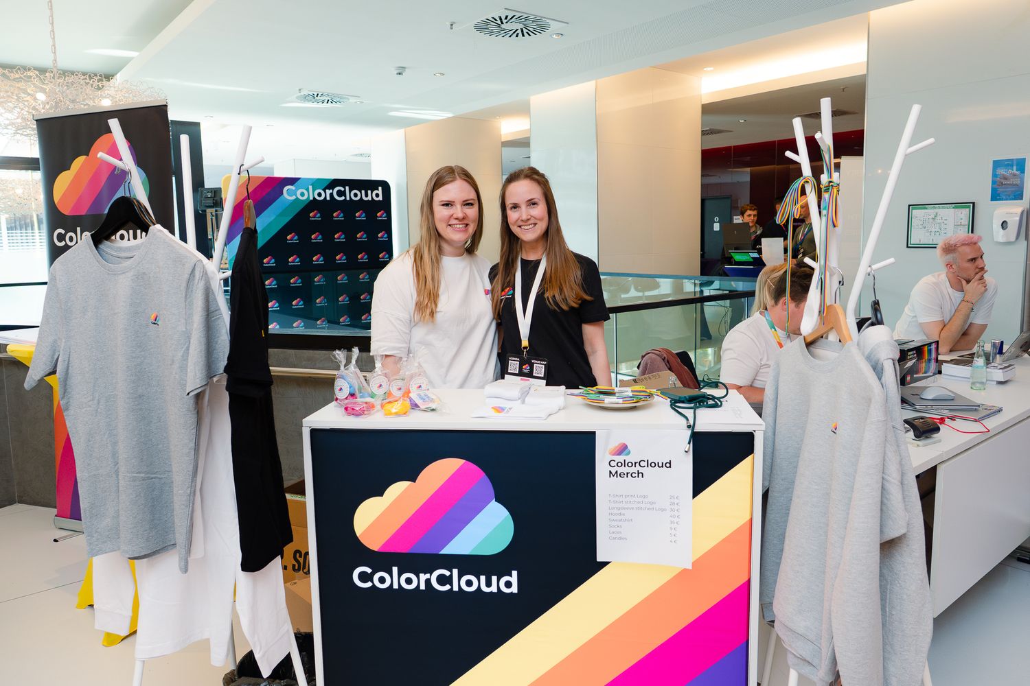 Paulina Meißner und eine weitere Mitorganisatorin  stehen hinter dem Merch-Stand der ColorCloud-Konferenz im Foyer und lächeln