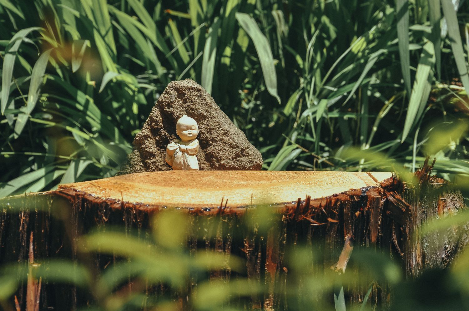 Eine kleine buddhistische Jizo-Statue auf einem Baumstumpf, umgeben von grünem Gras und Pflanzen
