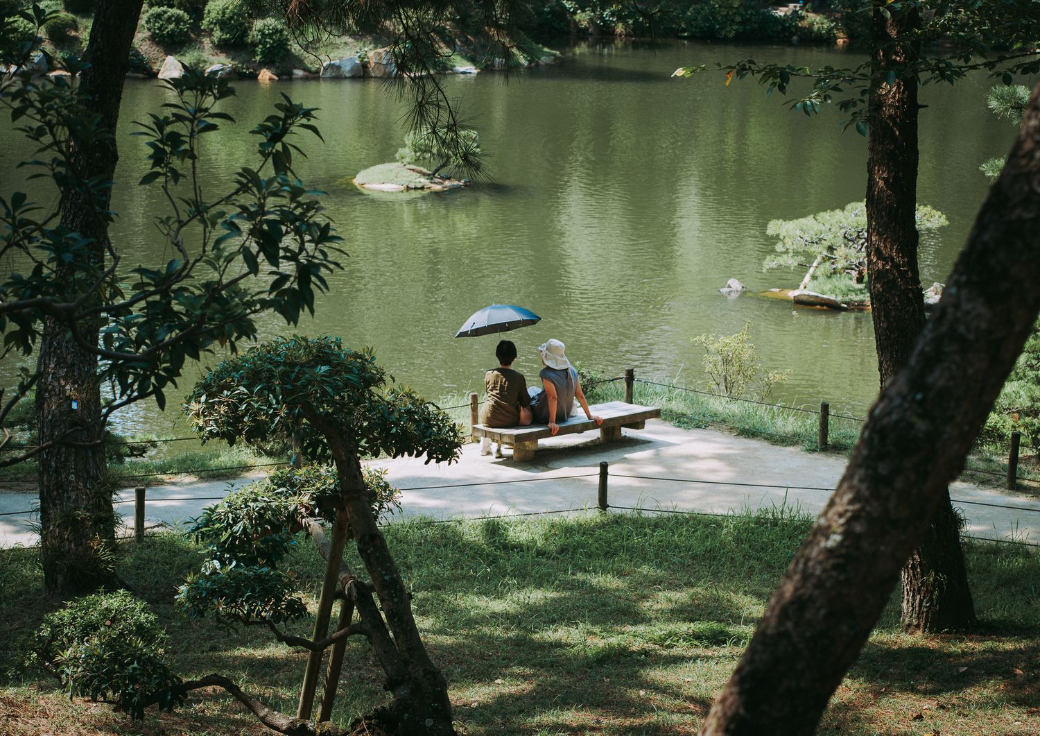 Zwei Frauen sitzen im Sonnenschein auf einer Bank in einem japanischen Garten; eine von ihnen hält einen blauen Sonnenschirm