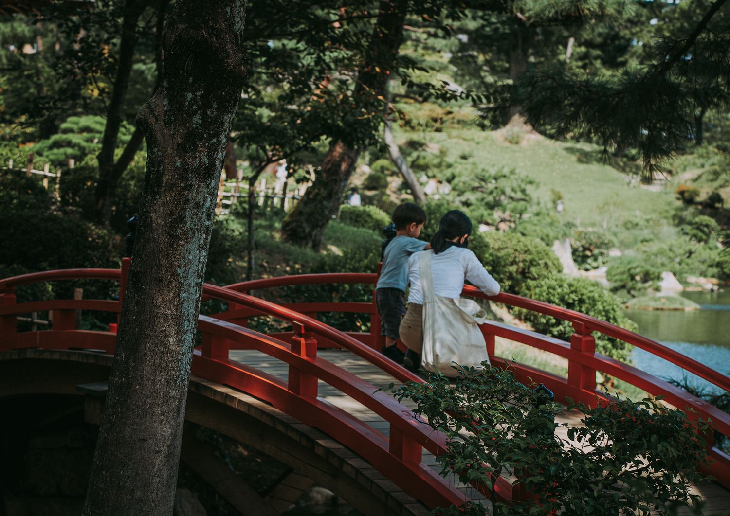 Eine Mutter steht mit ihrem Kind auf einer roten japanischen Holzbrücke in einem grünen Park und blicken auf einen Teich