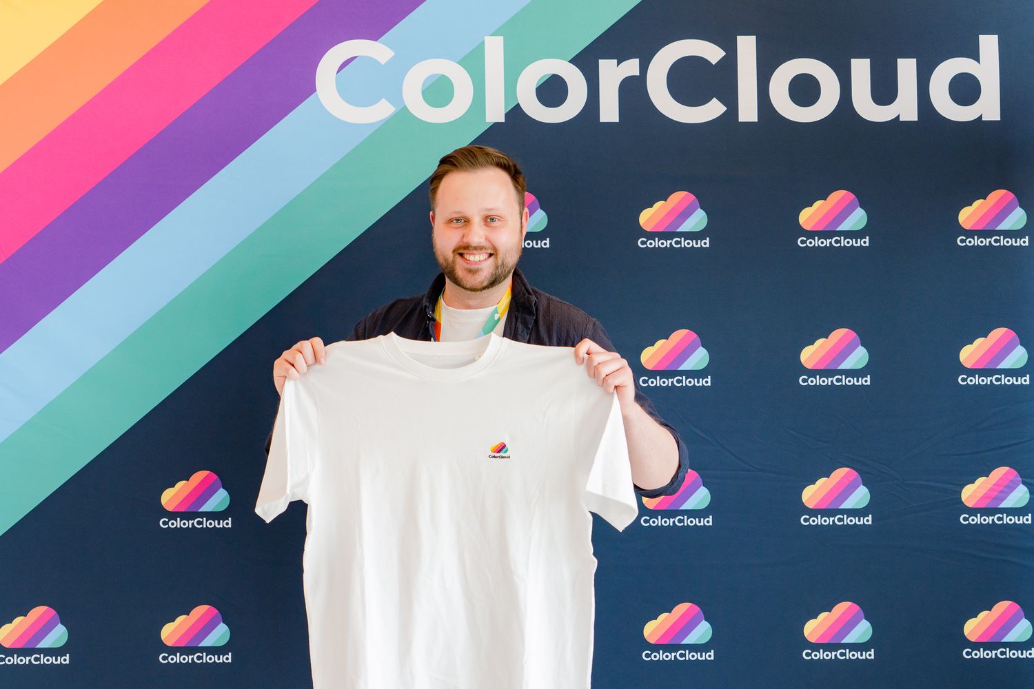Ein Teilnehmer posiert vor einer Fotowand und hält  mit beiden Händen ein weißes Merch-T-Shirt von der ColorCloud-Konferenz hoch