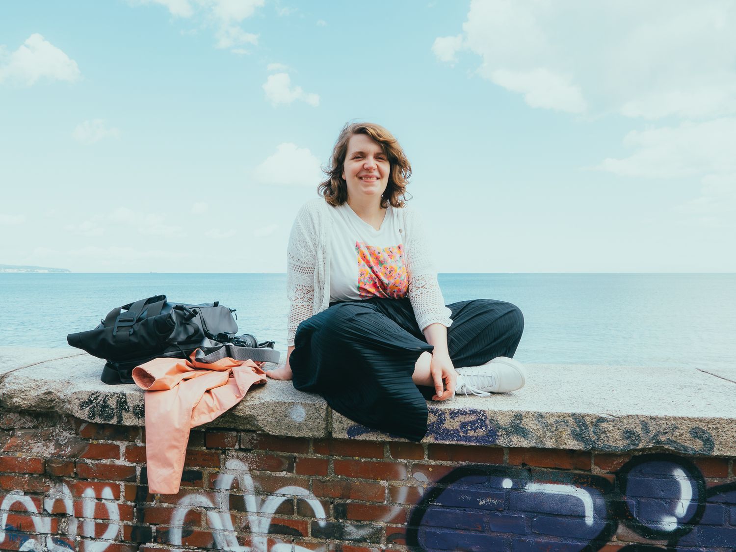 Fotografin Jana Spychalski sitzt im Schneidersitz auf einer Mauer, im Hintergrund ist das Meer
