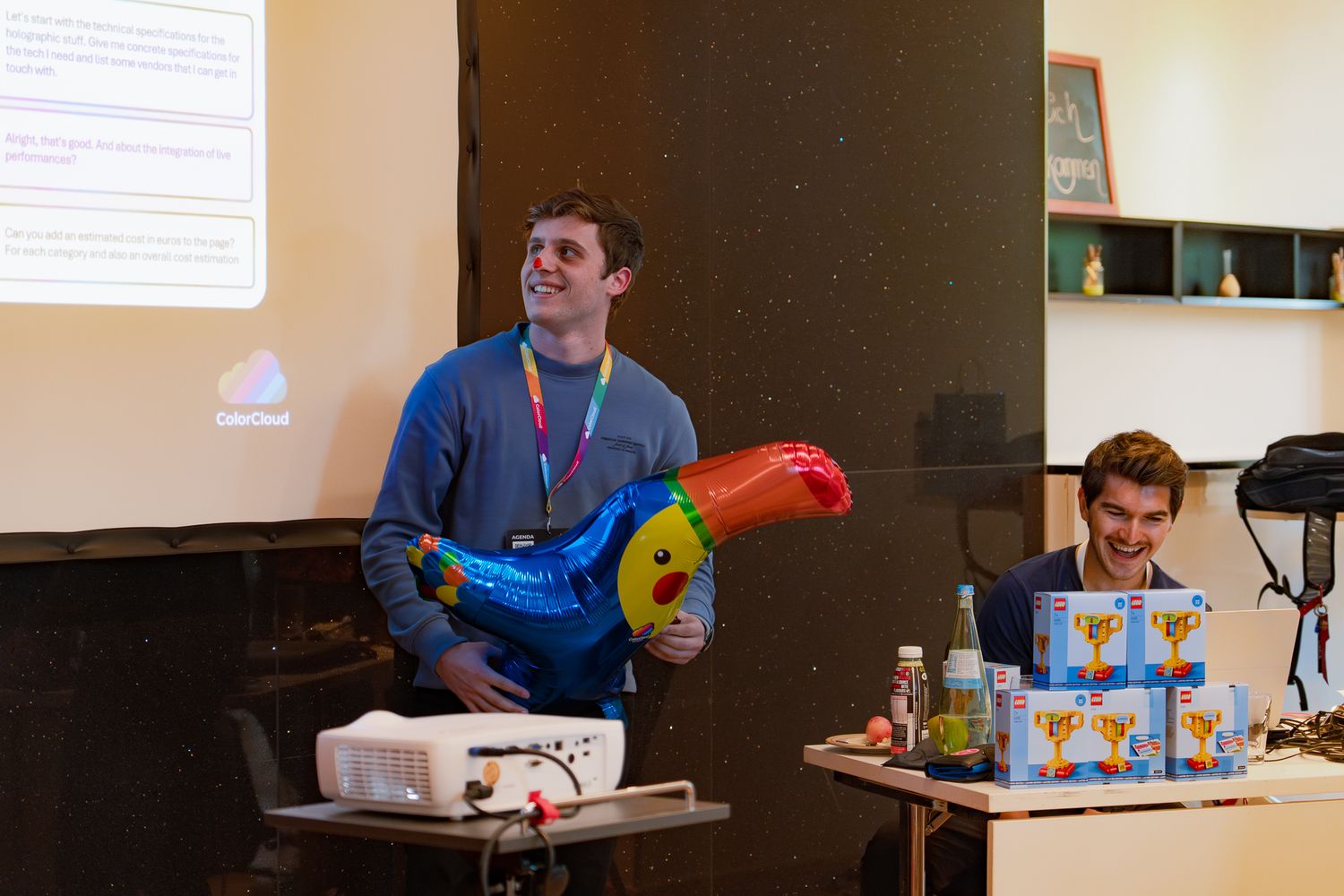 Ein Teilnehmer präsentiert beim ColorCloud PromptAthon Ergebnisse vor anderen Teilnehmenden und hält einen bunten Luftballon in der Hand