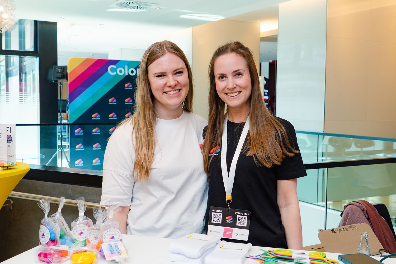 Paulina Meißner und eine weitere Mitorganisatorin  posieren hinter dem Merch-Stand der ColorCloud-Konferenz im Foyer und lächeln