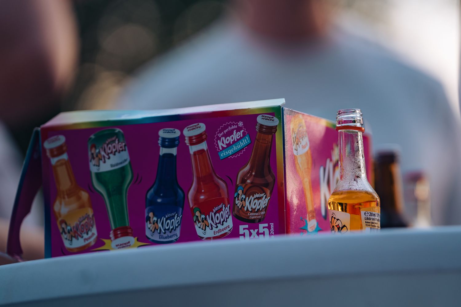 Verpackung der Marke ‚Kleiner Klopfer‘ mit bunten Likörfläschchen und einer geöffneten Flasche auf einem Tisch