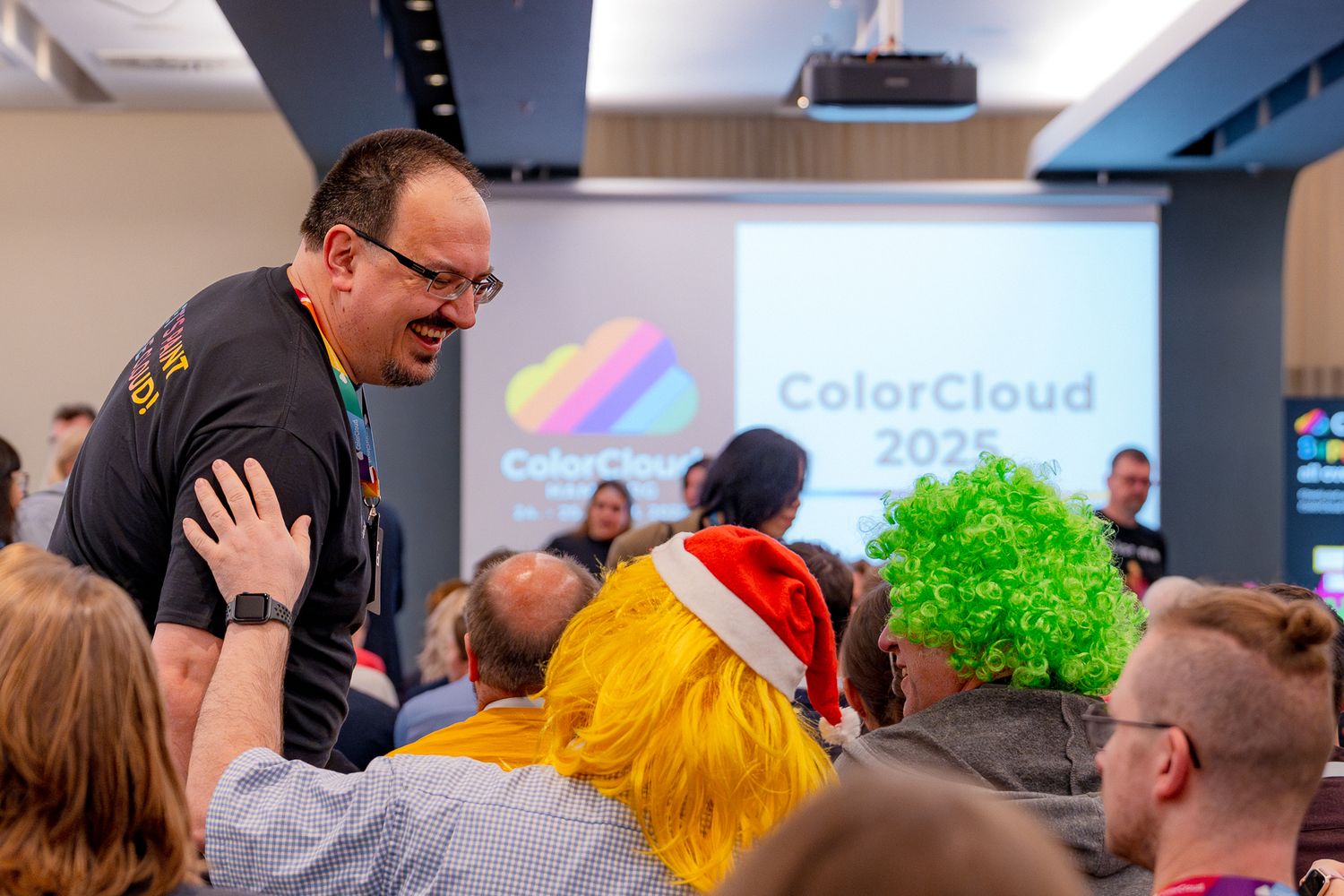 Ein Teilnehmer begrüßt zwei weitere Teilnehmer, die gelbe und grüne Perücken tragen, im Hintergrund steht 'ColorCloud 2025' auf der Leinwand