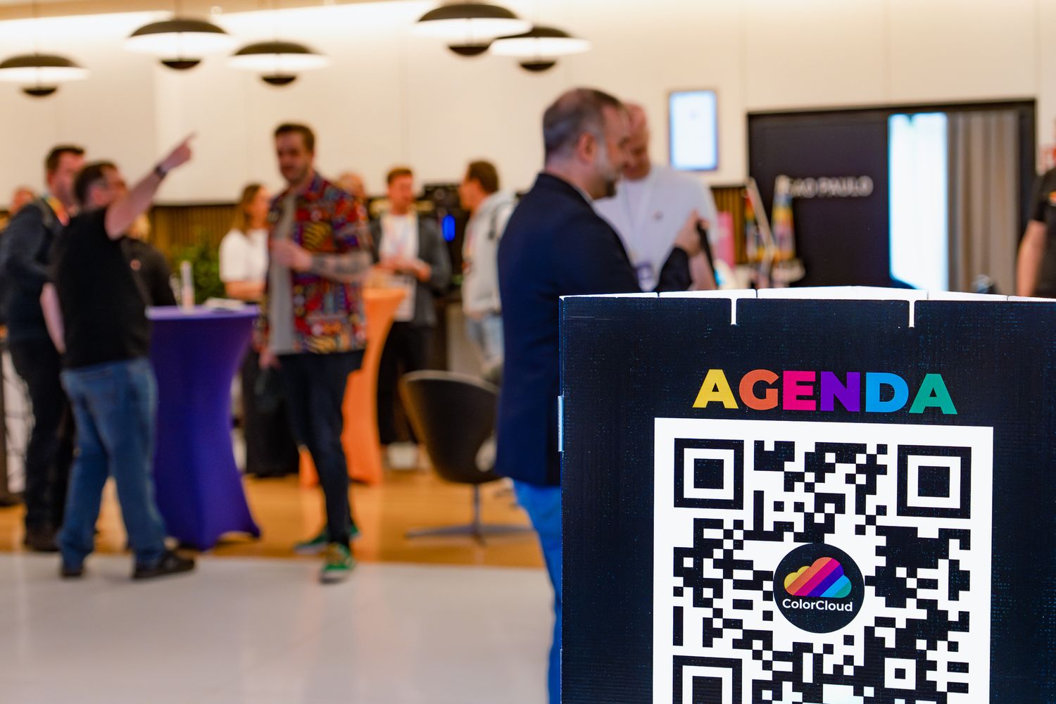 Teilnehmende an der ColorCloud unterhalten sich an Stehtischen im Foyer, im Vordergrund ist ein Pappaufsteller mit aufgedrucktem QR-Code