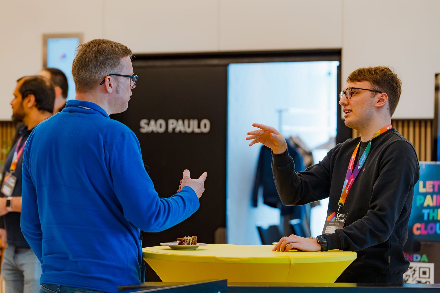 Zwei Teilnehmer der ColorCloud-Konferenz unterhalten sich mit den Händen gestikulierend an einem gelben Stehtisch im Foyer