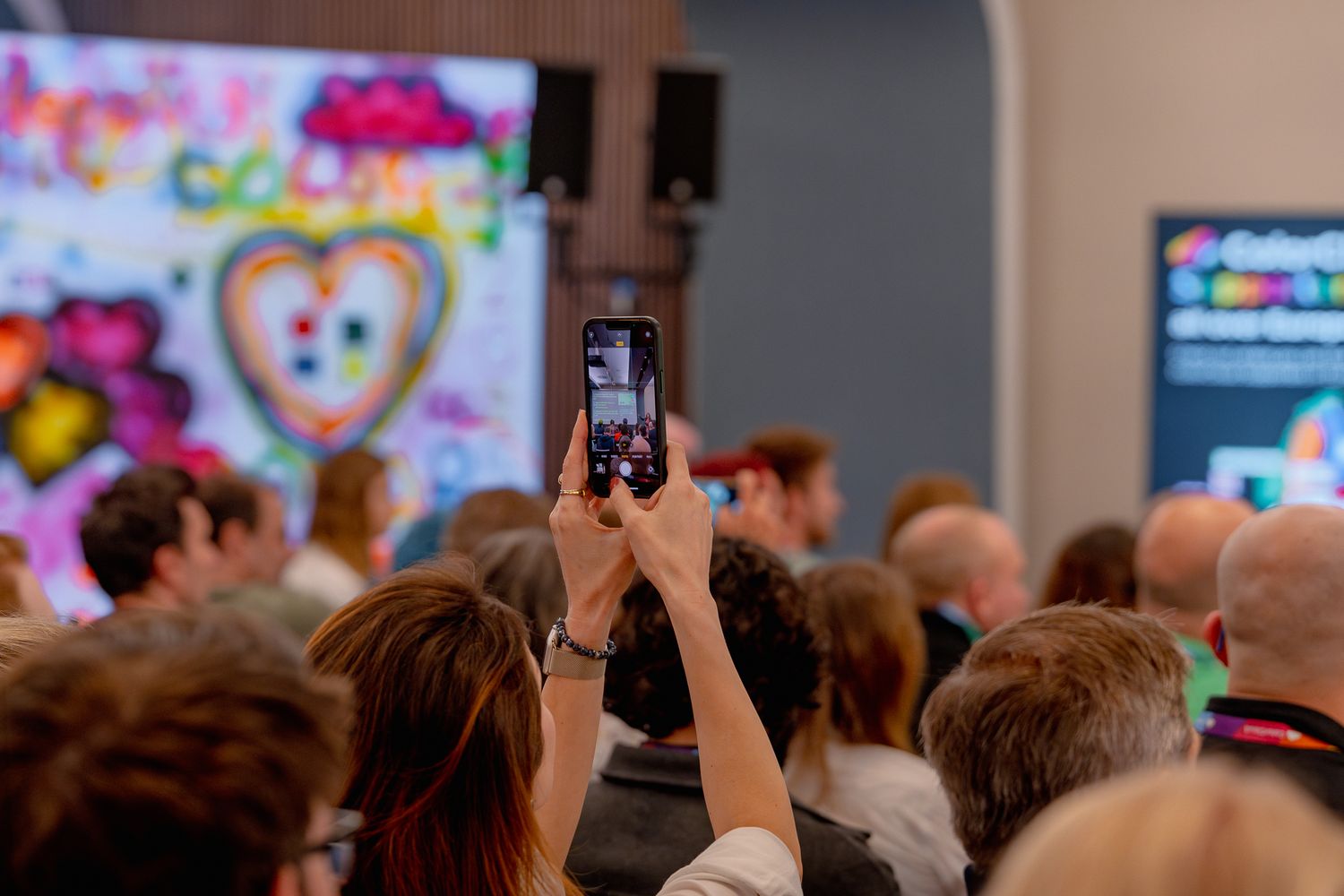 Eine Teilnehmerin aus dem Publikum fotografiert mit ihrem Handy während der Eröffnungsrede der ColorCloud 2025 das Geschehen