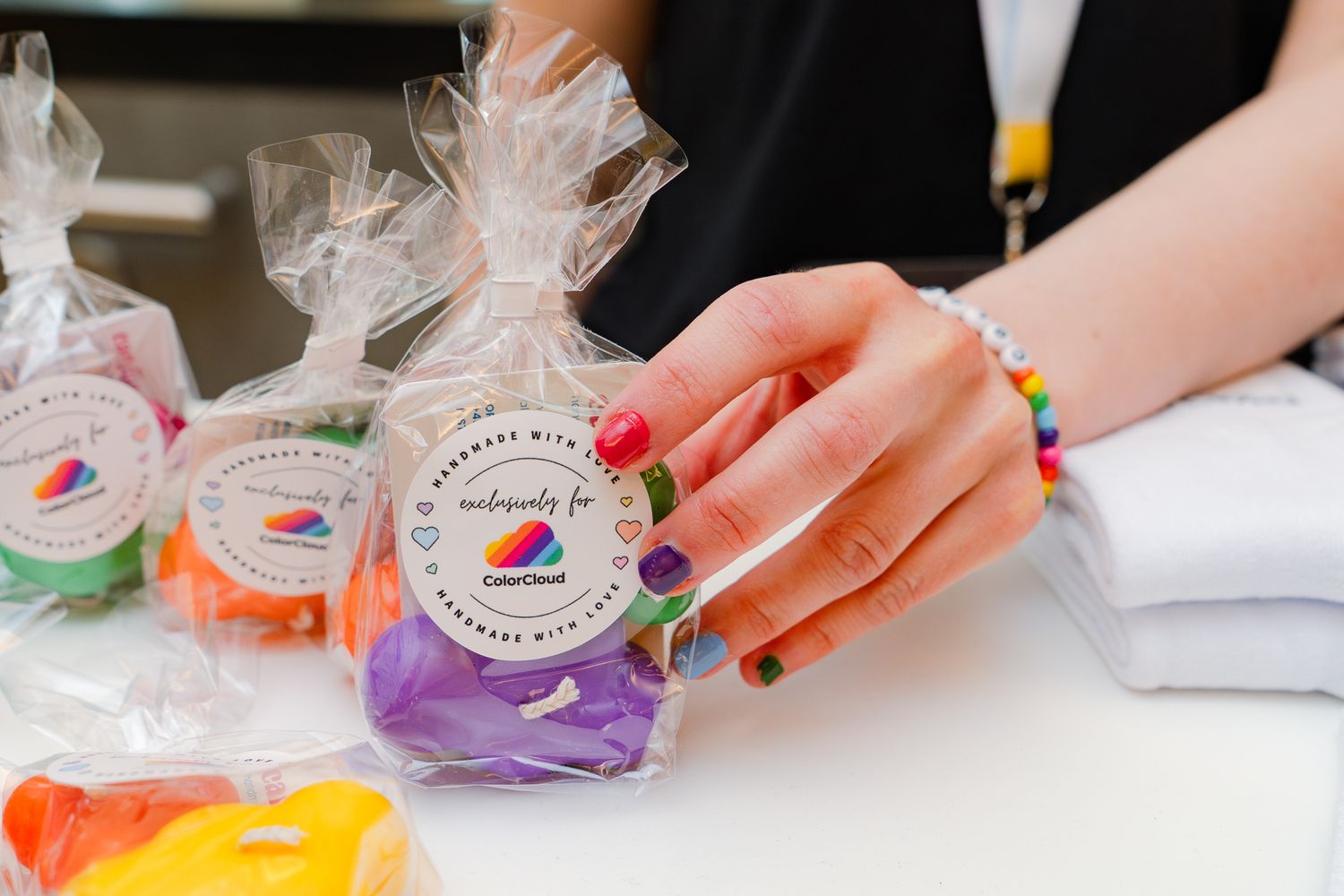 Eine Hand mit bunt lackierten Fingernägeln hält eine transparente Geschenktüte mit handgemachten lila Kerzen mit ColorCloud-Branding