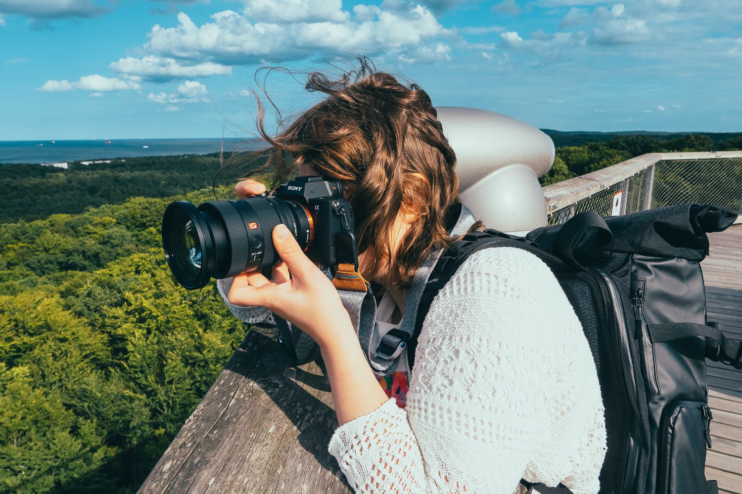 Fotografin Jana Spychalski lehnt sich an ein hölzernes Geländer und blickt durch den Sucher ihrer Sony-Kamera, im Hintergrund Wald und Meer