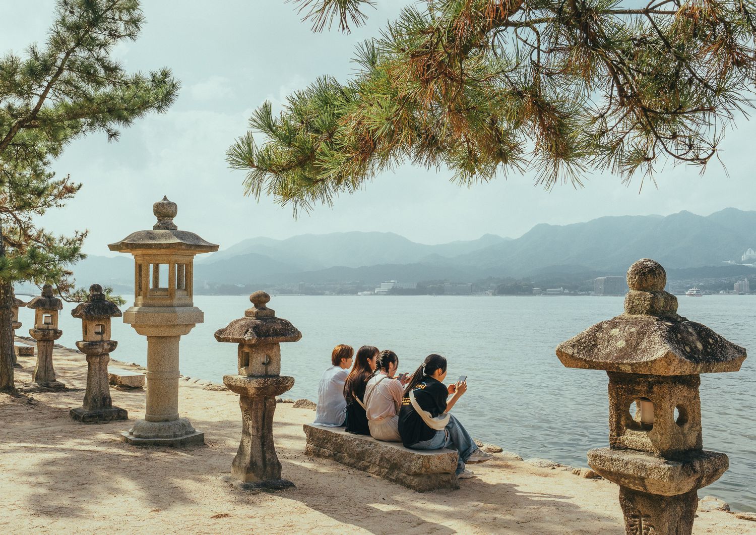 Vier Frauen sitzen auf einer steinernen Bank an der Küste der Insel Miyajima; neben ihnen stehen traditionelle japanische Steinlaternen.