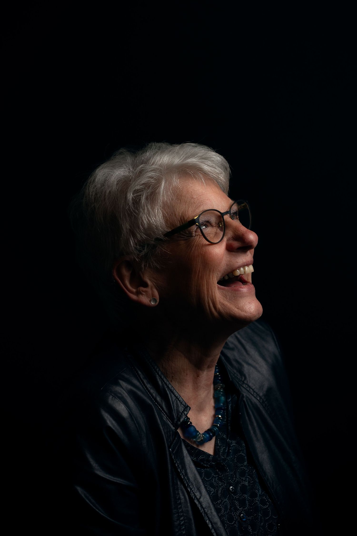 Portrait einer älteren Frau im Profil mit weißen Haaren, sie hat den Kopf leicht nach oben zum Licht geneigt und lacht breit